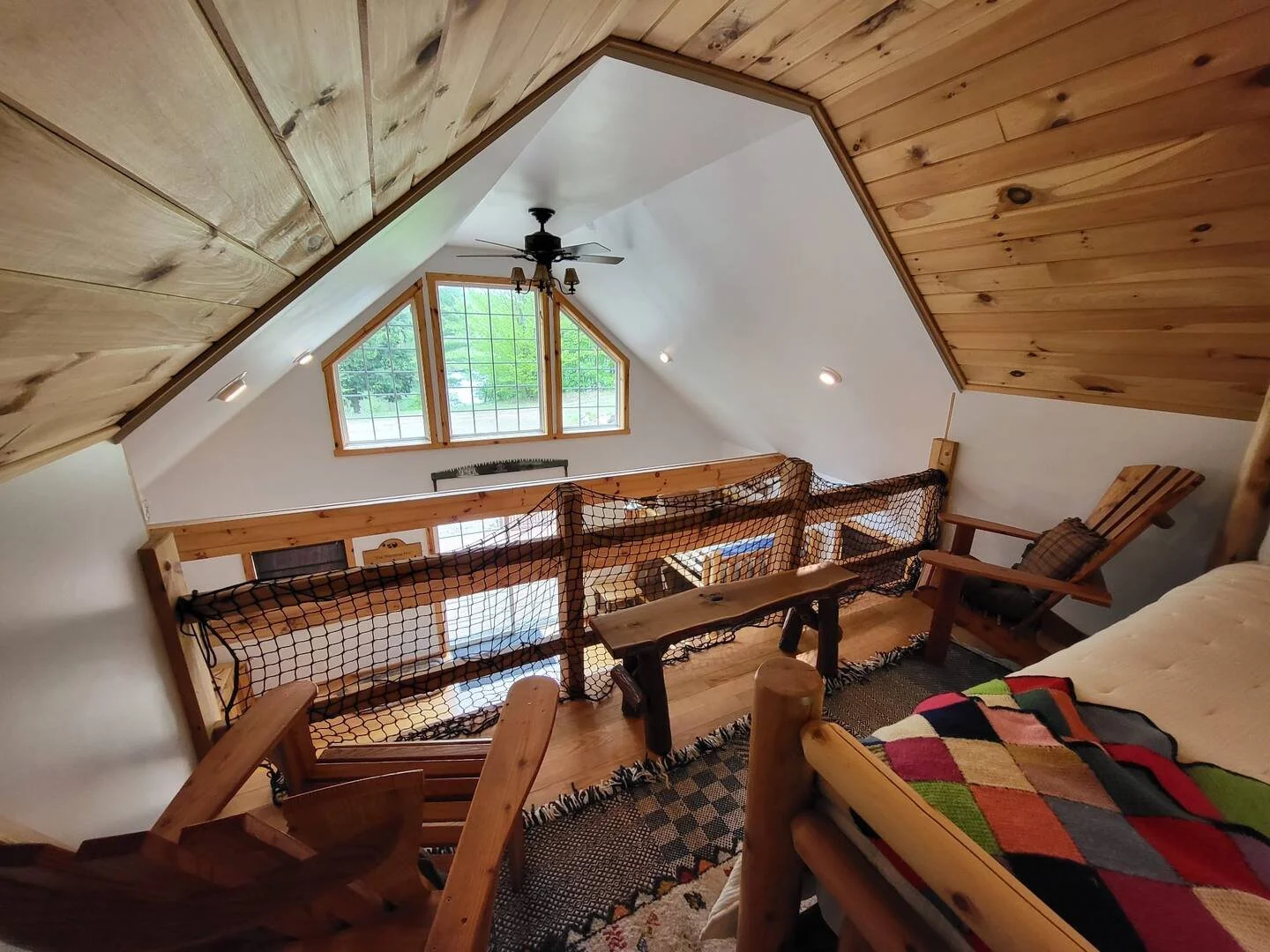 Interior of a cozy loft bedroom with wood-paneled ceiling, a ceiling fan, large window, and rustic furniture including a bed with a colorful quilt and a rocking chair.
