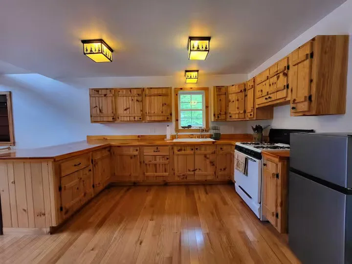 Kitchen with wooden cabinets, white stove, gray refrigerator, small window above the sink, and wooden floors.