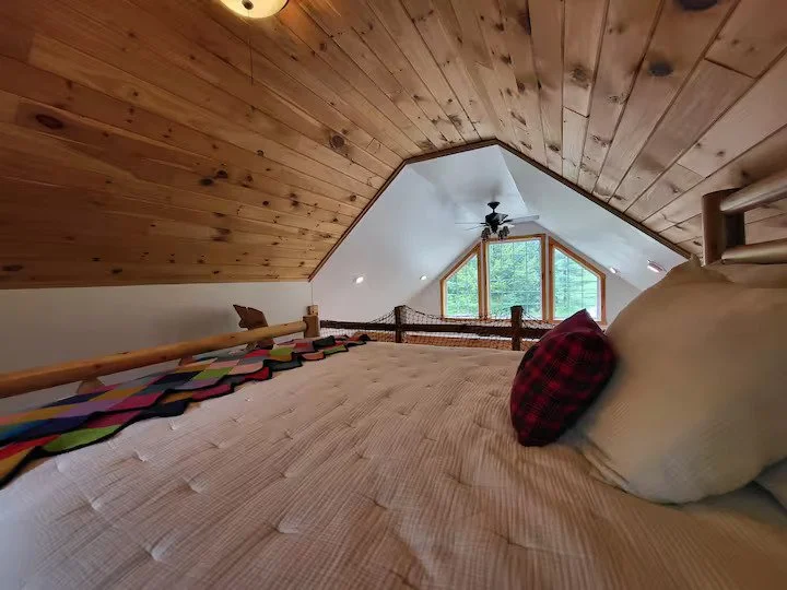View of a cozy attic bedroom with a sloped wooden ceiling, a large window, a ceiling fan, and a bed with pillows in the foreground.