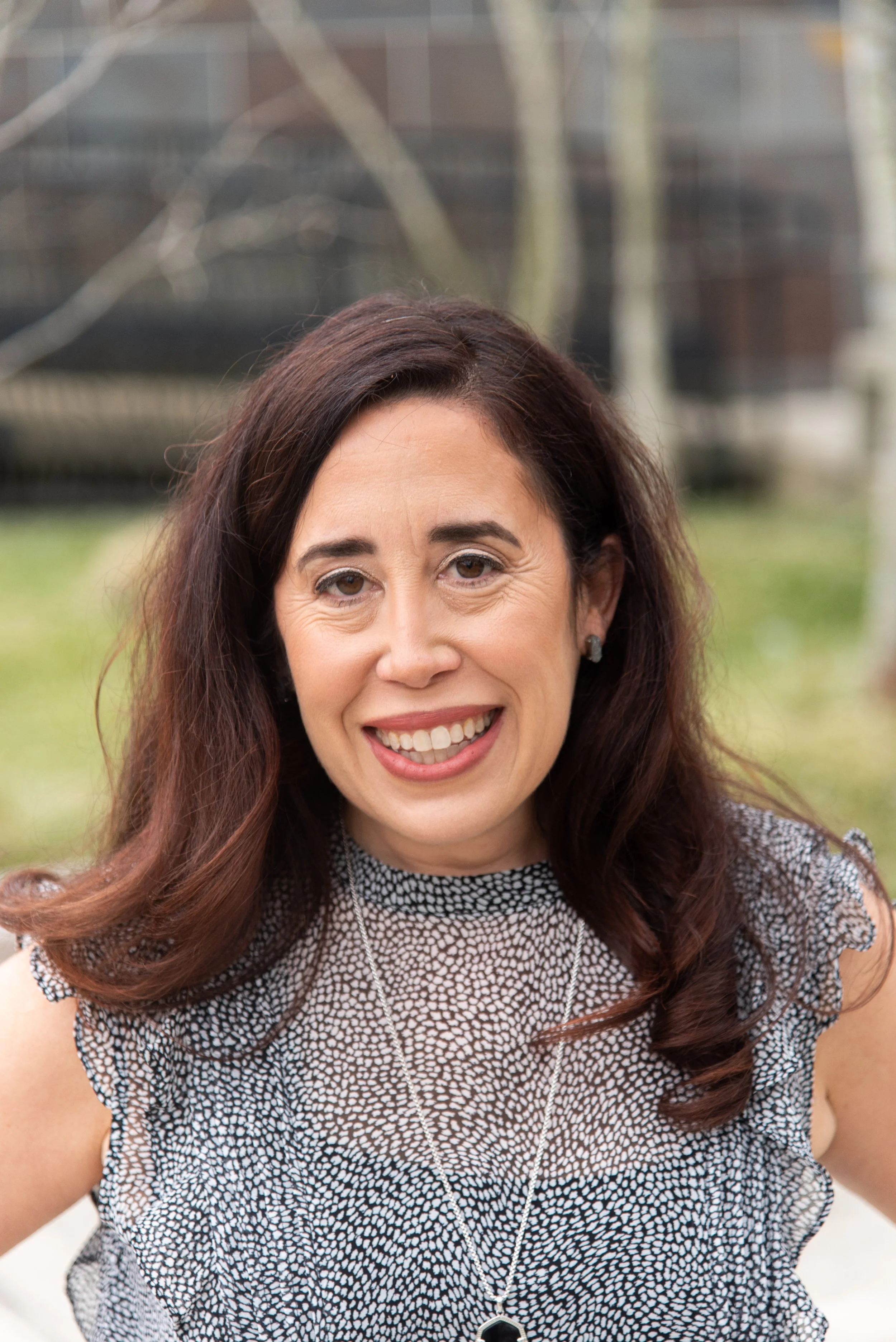 A smiling woman with shoulder-length brown hair, wearing a patterned sleeveless top and a necklace, outdoors with a blurred background of trees and buildings.