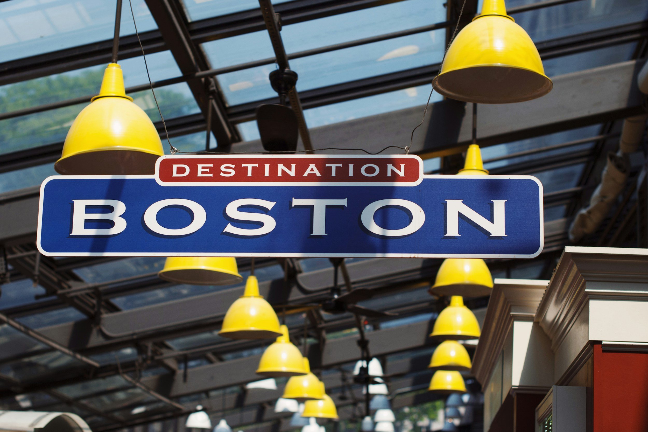 Sign that reads 'Destination Boston' hanging above yellow pendant lights inside a glass-roofed structure.