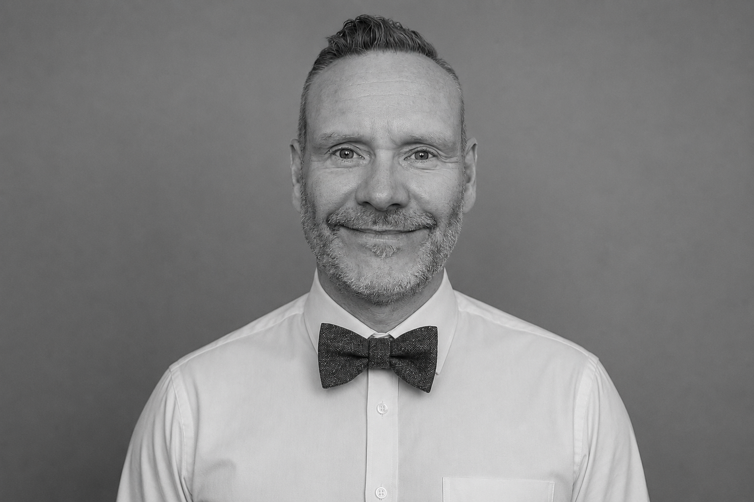 Black and white photograph of a smiling man with a beard, wearing a white shirt and a bow tie, against a plain background.