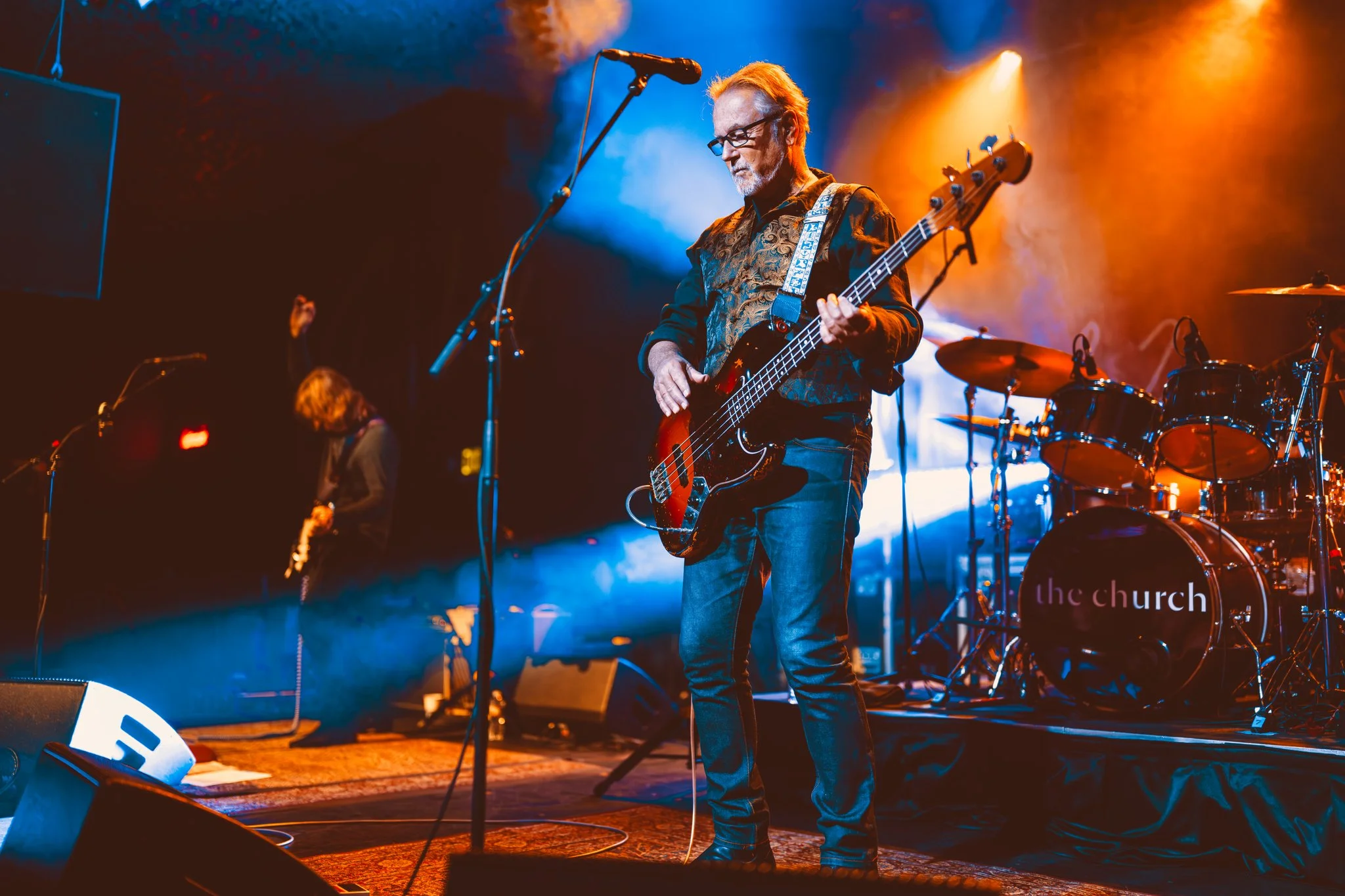 A man with glasses and a beard playing a bass guitar on stage under colorful lights, with drums in the background, during a concert or music performance.