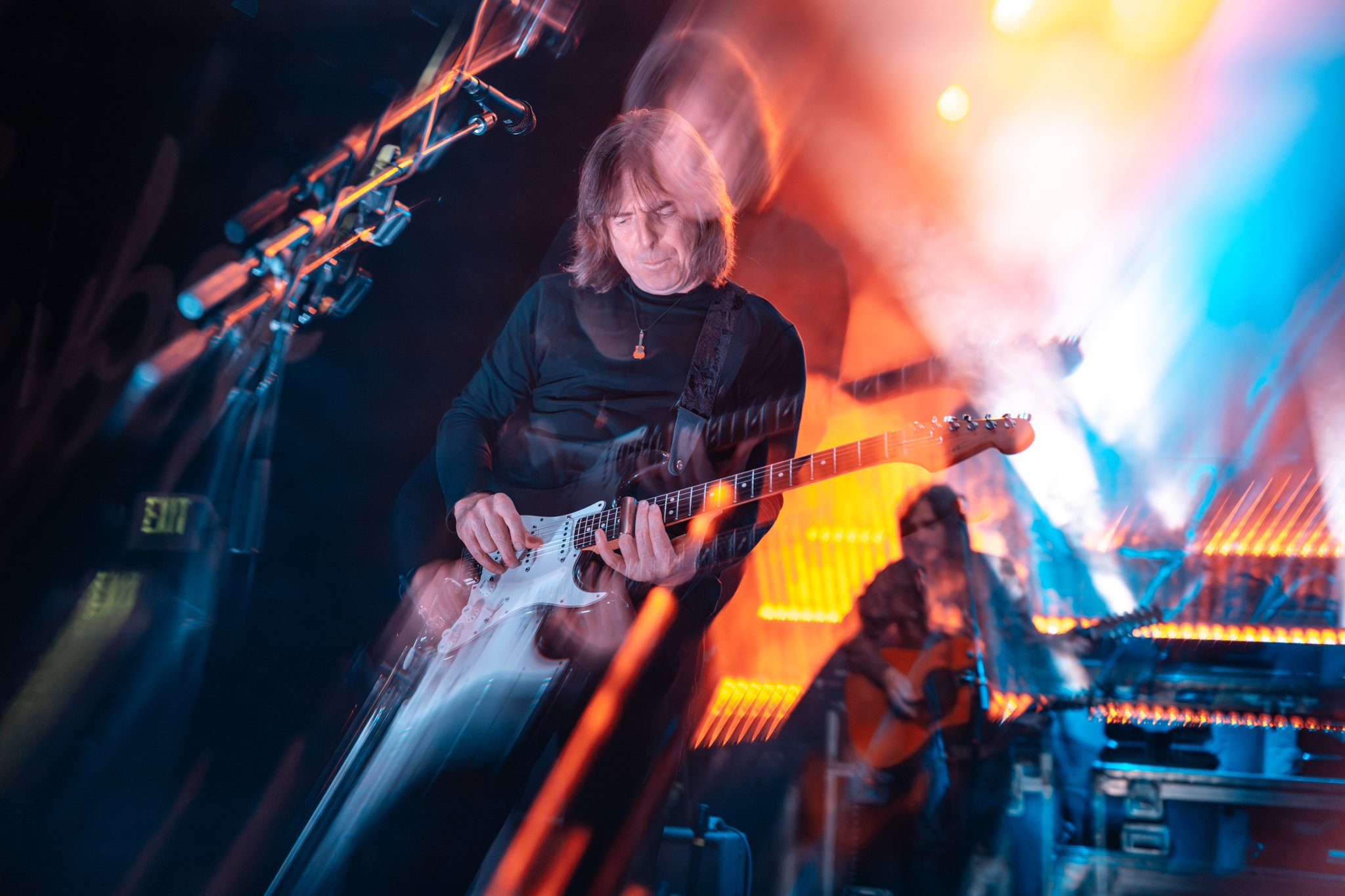 A man with long hair playing an electric guitar on a stage with colorful stage lights and smoke effects.