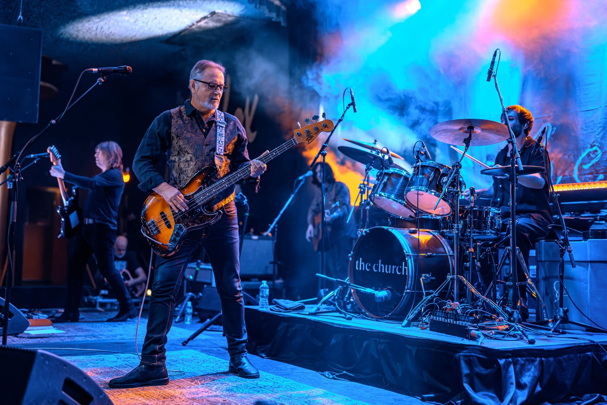 A band performing on stage with a male bassist in the foreground, a drummer behind him, and colorful stage lighting creating a vibrant atmosphere.