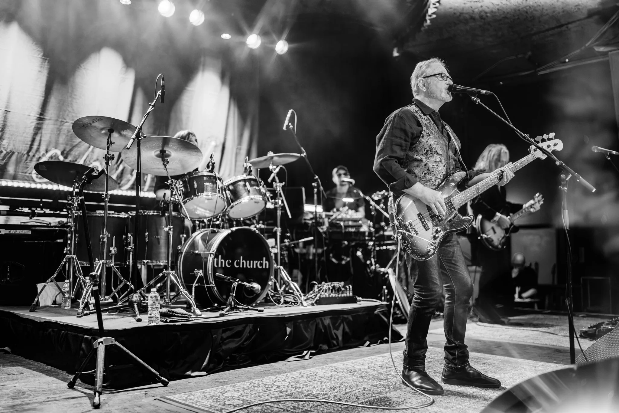 Black and white photo of a live band performing on stage. A man in glasses, a vest, and jeans is playing a bass guitar and singing into a microphone. Behind him, there is a drum set labeled "the church," and other band members with instruments are vi