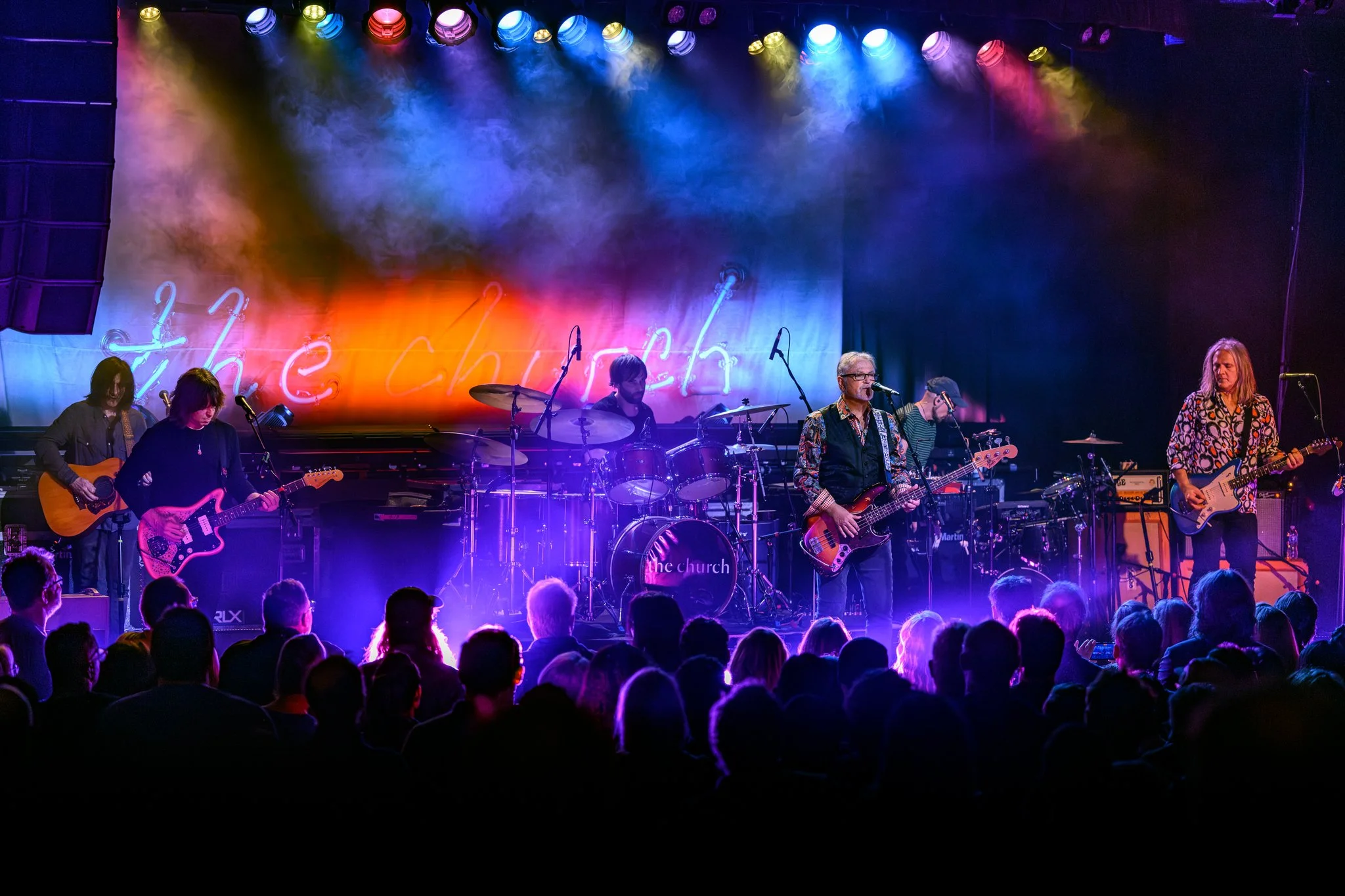 A band performing live on stage with colorful lighting and a neon sign that says 'the church' in the background, while audience members watch.