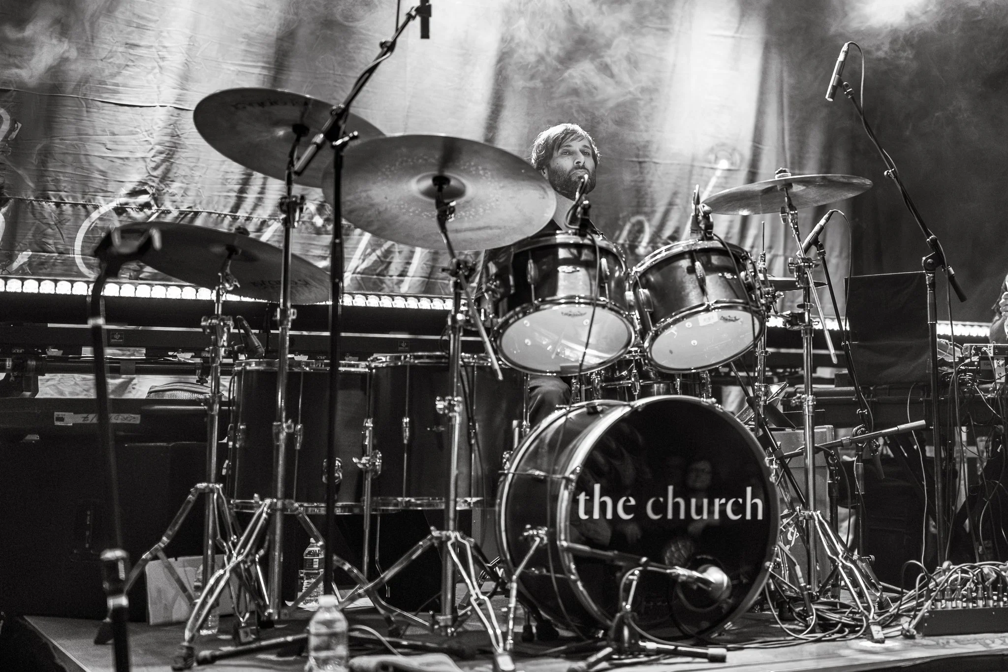 Black and white photo of a drummer playing a drum set on stage, with microphones positioned around the drums and a backdrop with the words "the church" partially visible.