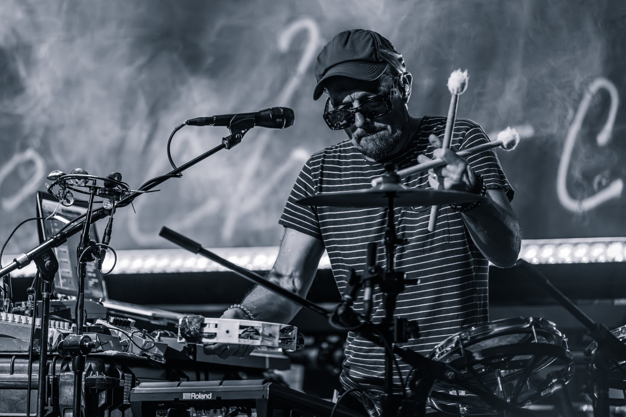 A man with a gray beard, wearing a cap, sunglasses, and a striped shirt, adjusting electronic music equipment and a microphone in a performance setting.