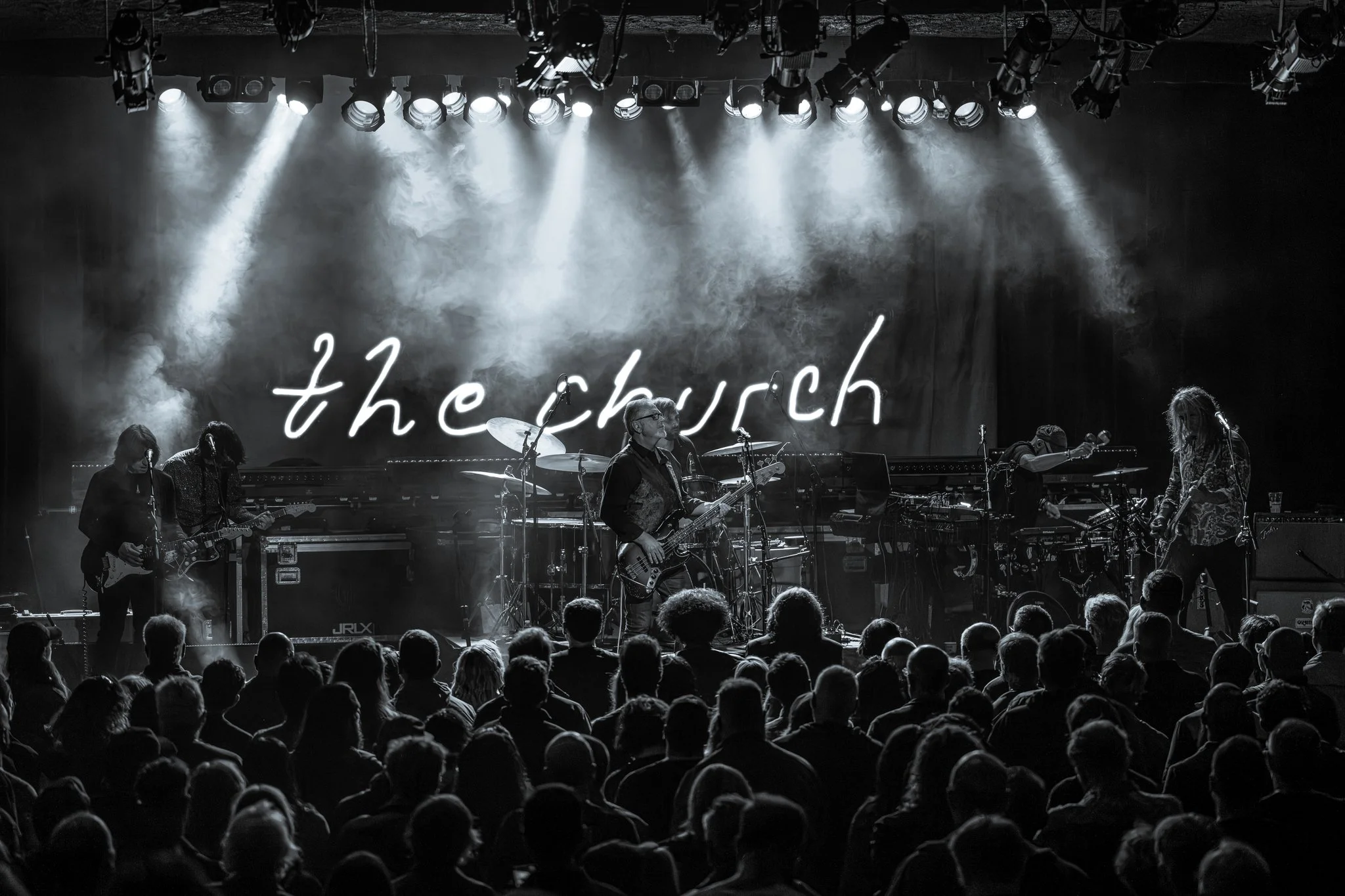 A live musical performance on stage with band members playing guitars, drums, and keyboard. Behind them, a neon sign reads 'the church'. The stage is lit with multiple spotlights, and an audience is visible in front of the stage.