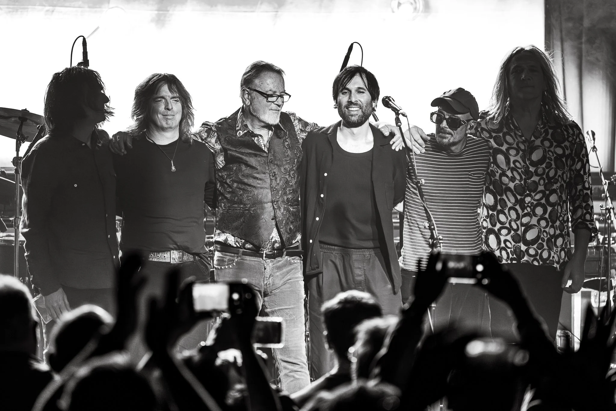 Black and white photograph of six male musicians standing side by side on stage, arm in arm, with instruments and microphones in the background, facing an audience.