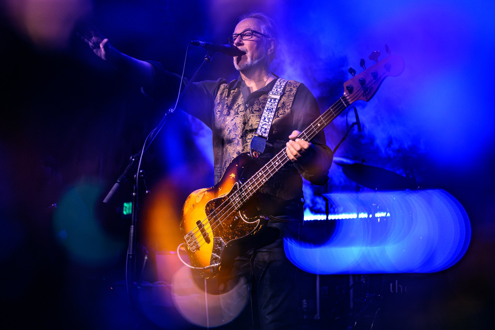 A man with glasses and a beard playing an electric bass guitar on stage, illuminated by blue and purple stage lights, with fog and microphone in front of him.