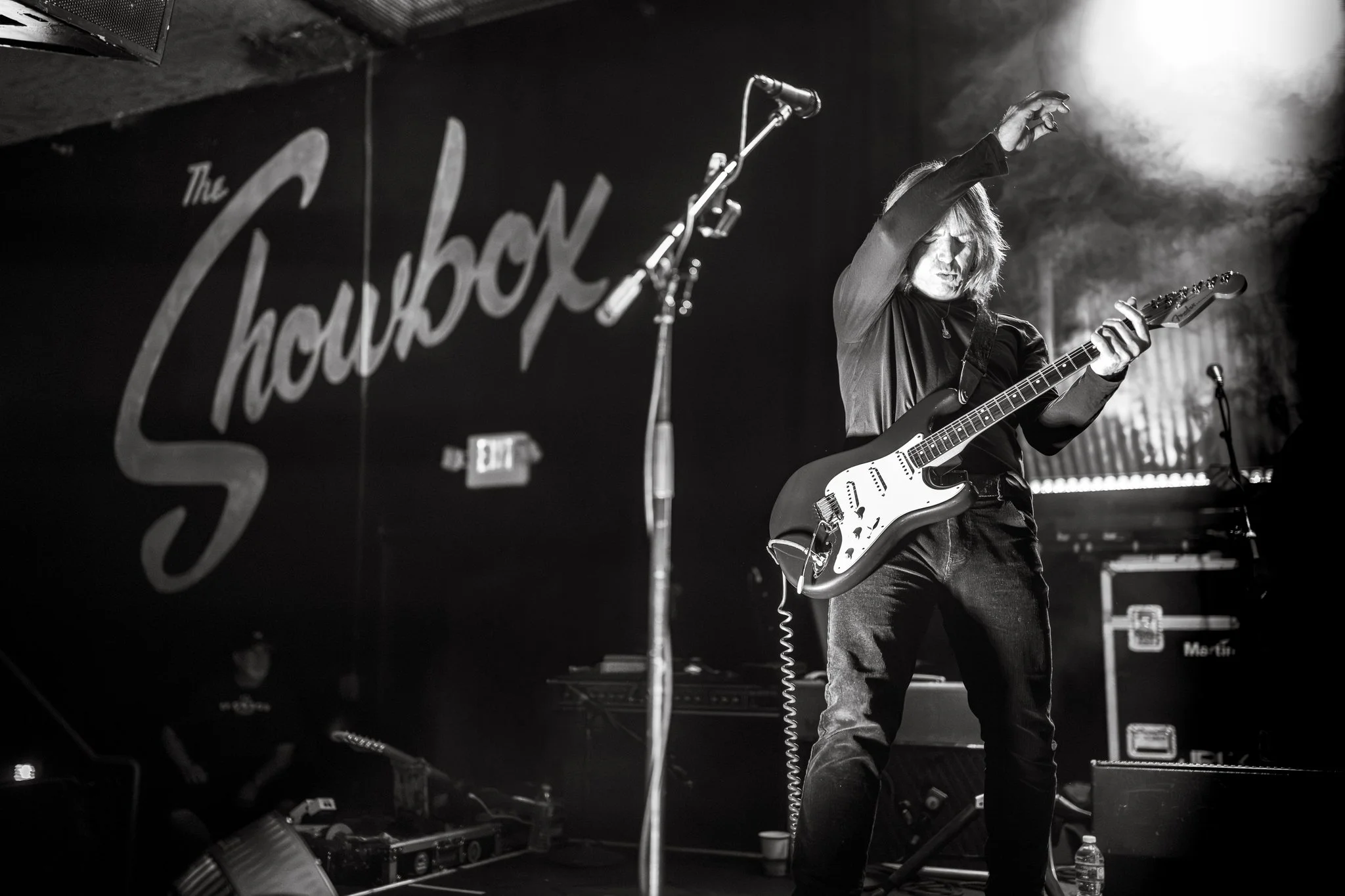 A musician playing an electric guitar on stage at a venue called The ShowBox The photo is in black and white and captures the musician from the side, with long hair and a dark shirt. There is stage equipment and a microphone in front of the musician.