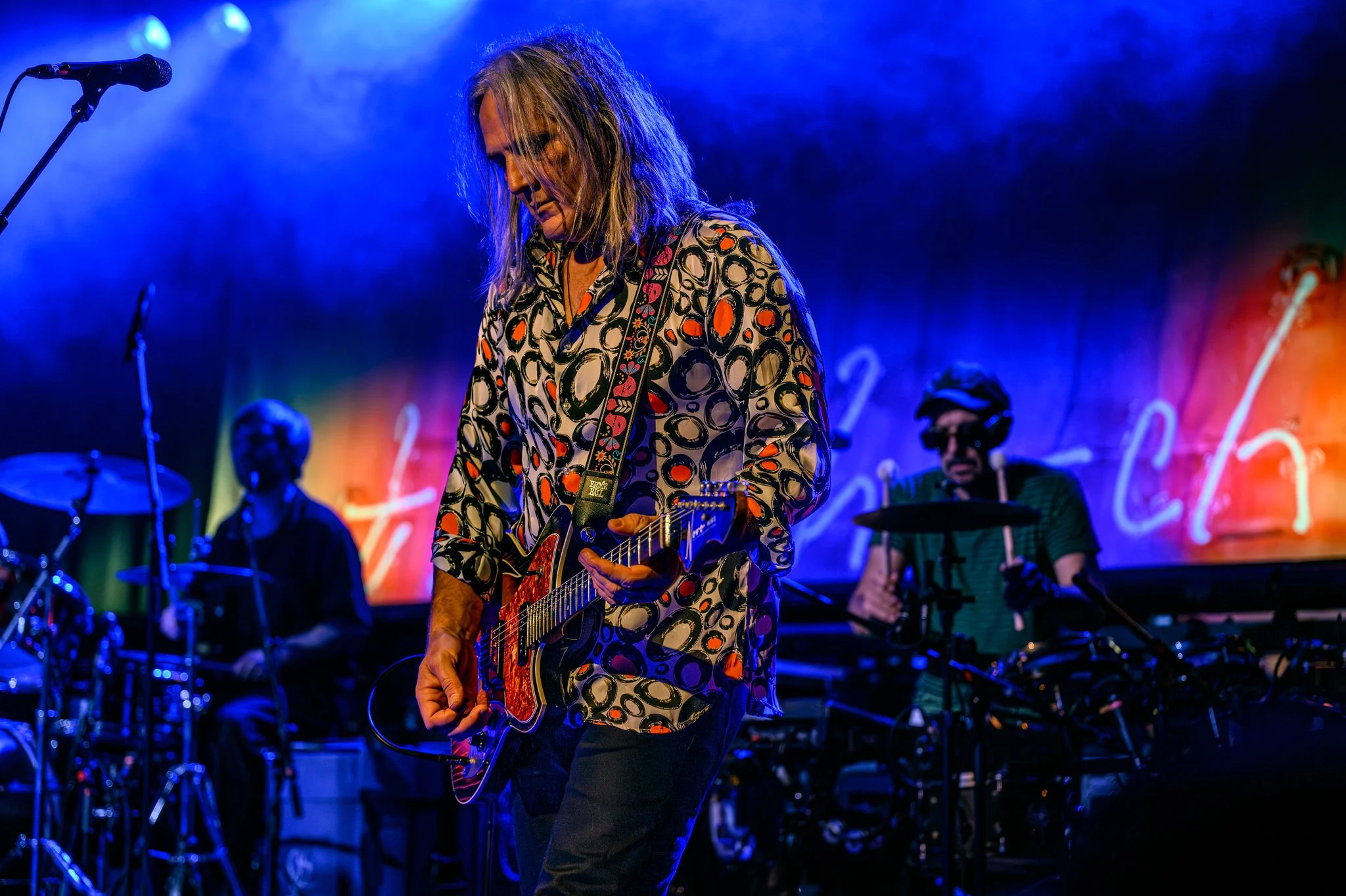 A musician with long hair playing an electric guitar on stage with vibrant colorful lighting and band members in the background.