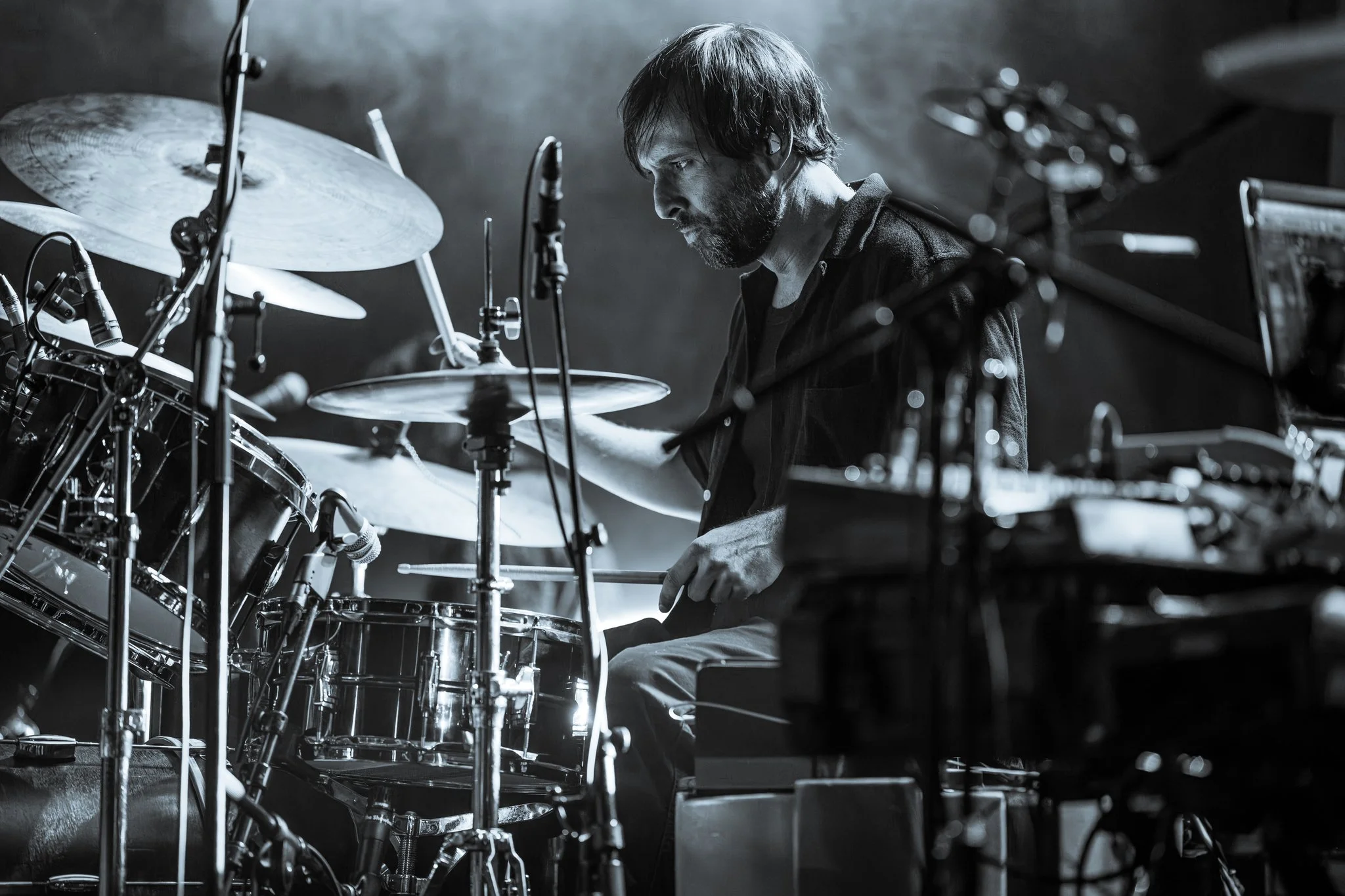 A man playing drums on stage, seen in profile, with intense focus. The image is in black and white, highlighting the scene's mood and atmosphere.