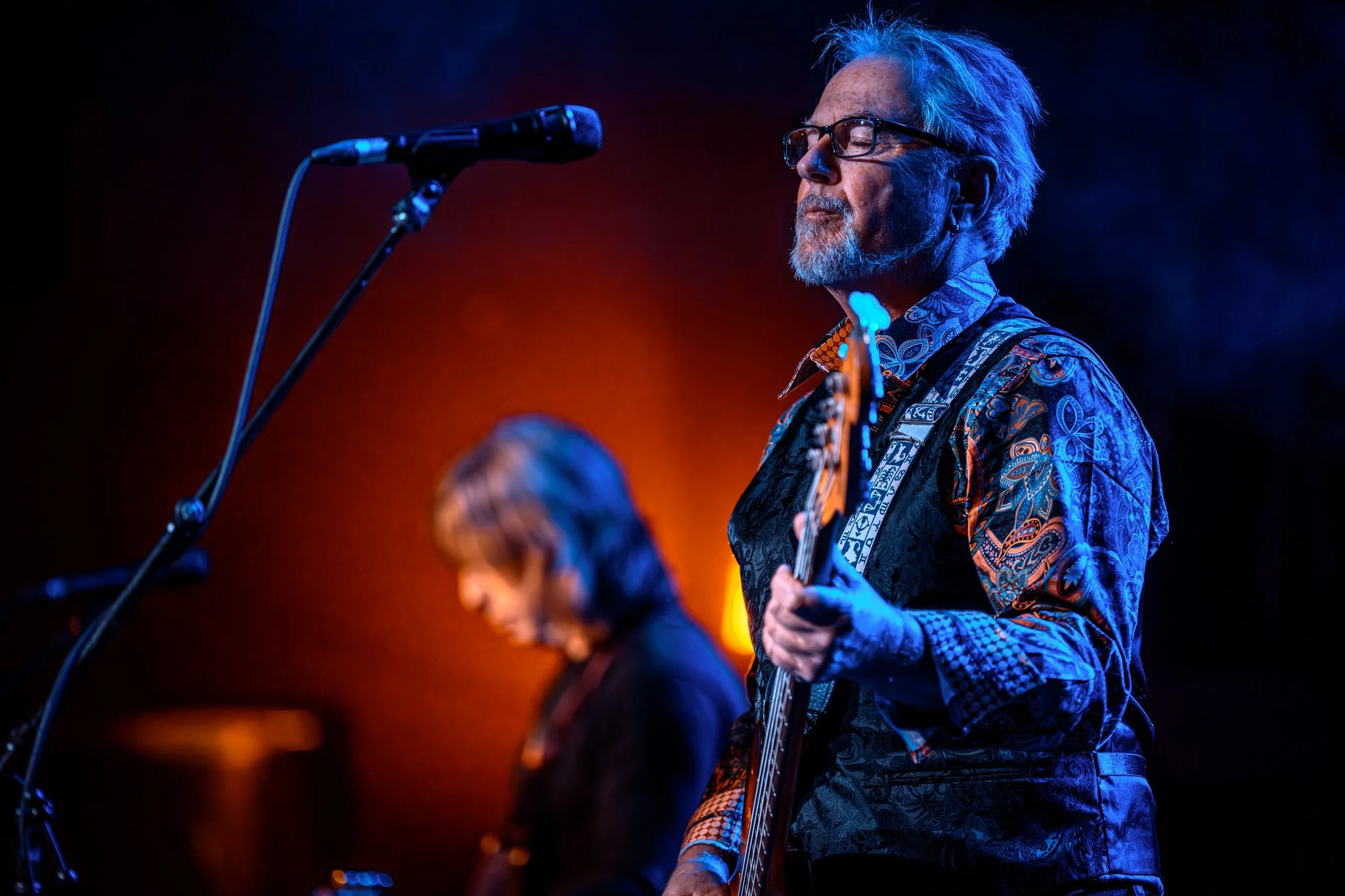 An elderly man with glasses and a white beard, dressed in a colorful patterned shirt, is playing an electric guitar on stage. His eyes are closed, and he appears to be deeply immersed in the music. There is a microphone in front of him, and the backg