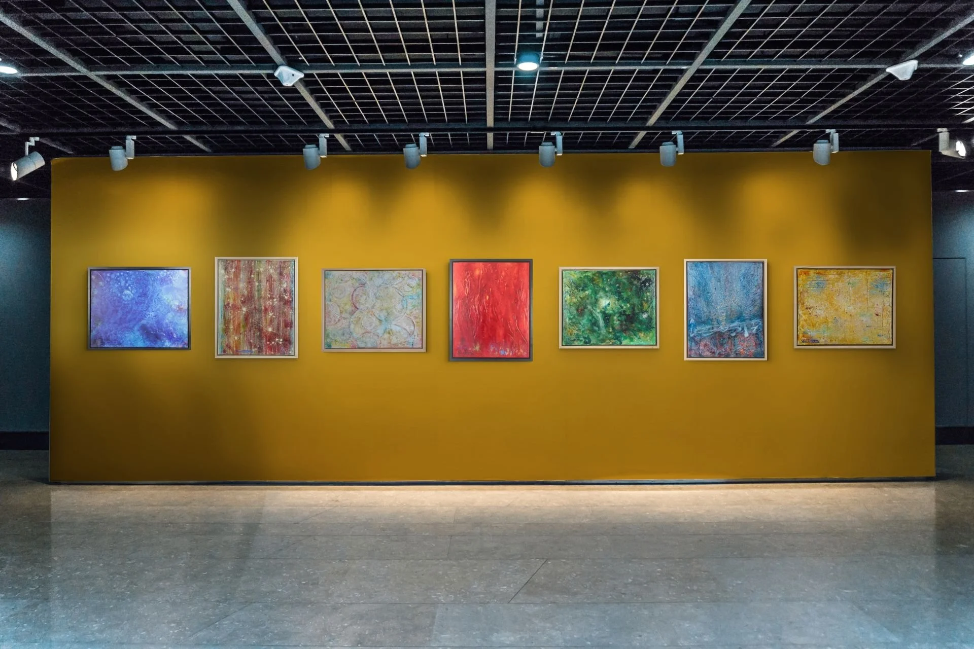 Six contemporary abstract paintings by Artist Shanti Padme, displayed on a deep yellow gallery wall, illuminated by ceiling track lights in an art gallery.