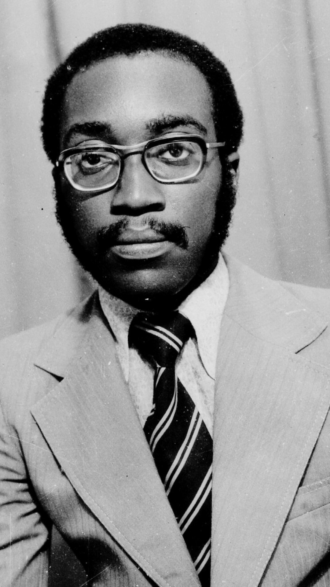 A black-and-white portrait of a man with glasses, a mustache, and beard, wearing a light-colored suit, a patterned shirt, and a dark striped tie, looking directly at the camera.