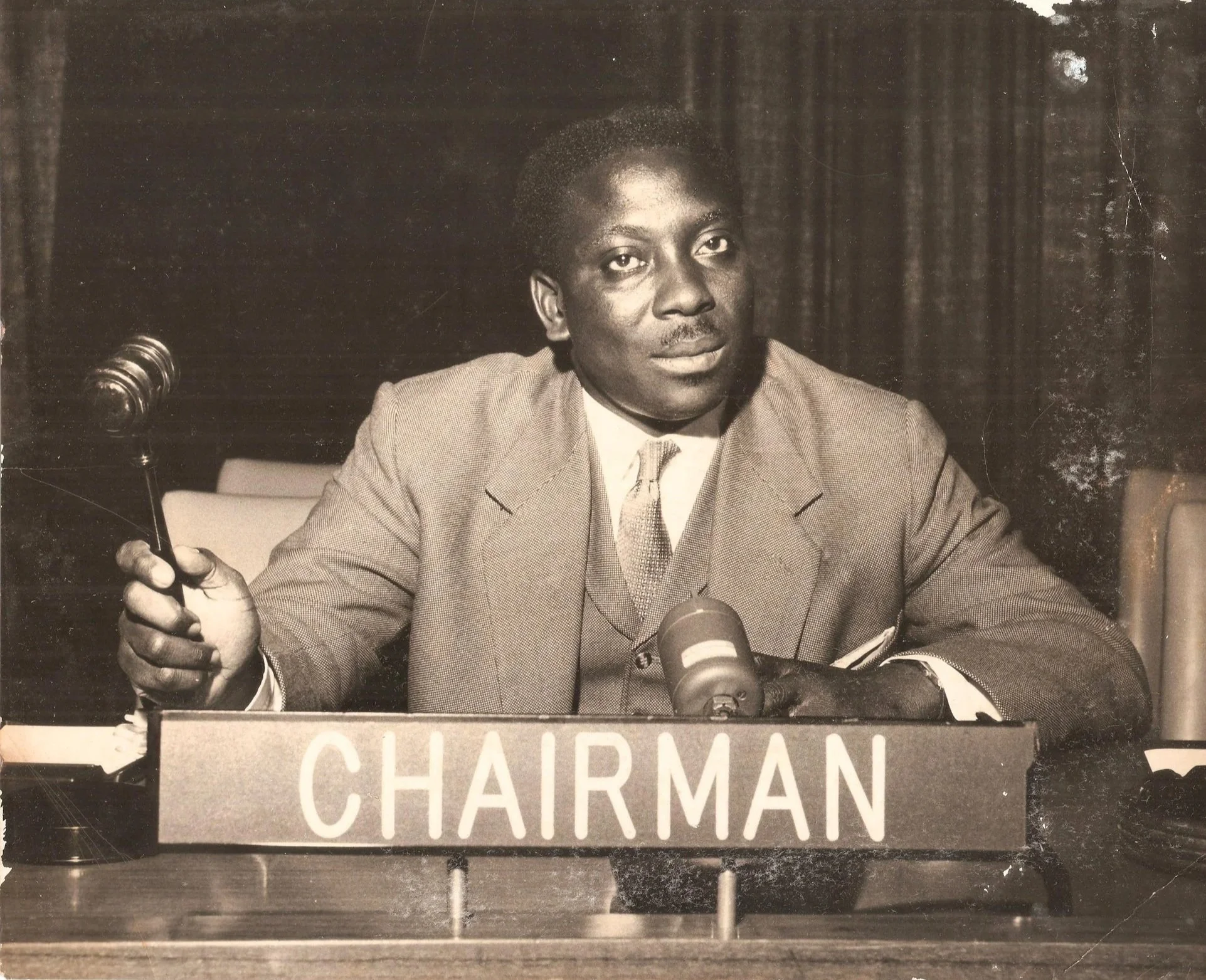 A man in a suit and tie sitting at a desk in front of a microphone with a sign that reads 'Chairman'.
