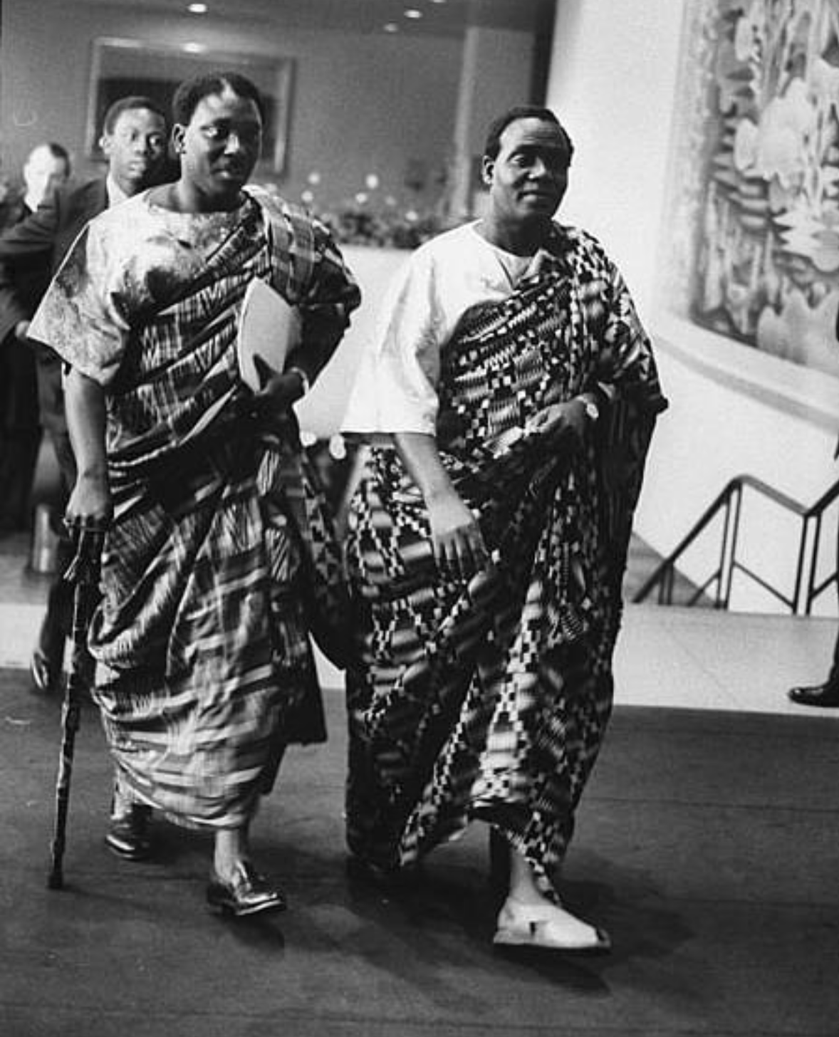 Two men dressed in traditional African attire, walking inside a gallery or museum, with artworks hanging on the wall in the background.