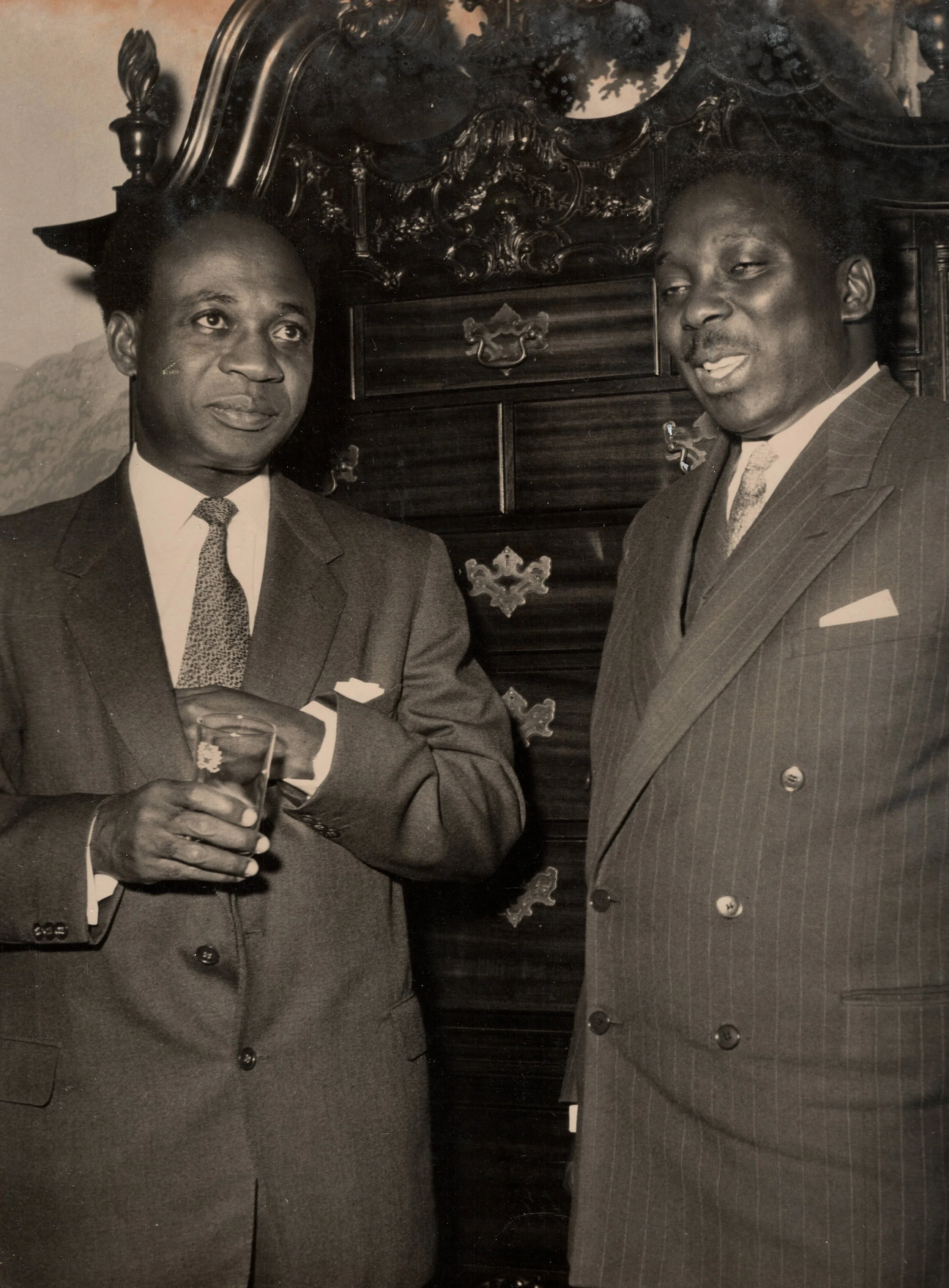 Two men in suits standing next to each other in a formal setting, with a dark ornamental wooden piece behind them. One man has a drink in his hand, and they are engaged in conversation.