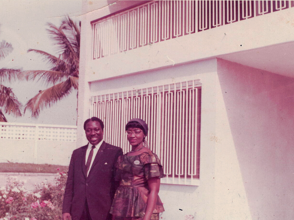 A man and woman standing outside a building with metal bars on the windows. The man is wearing a dark suit, and the woman is wearing a colorful dress with glasses and a headband. Palm trees are visible in the background.