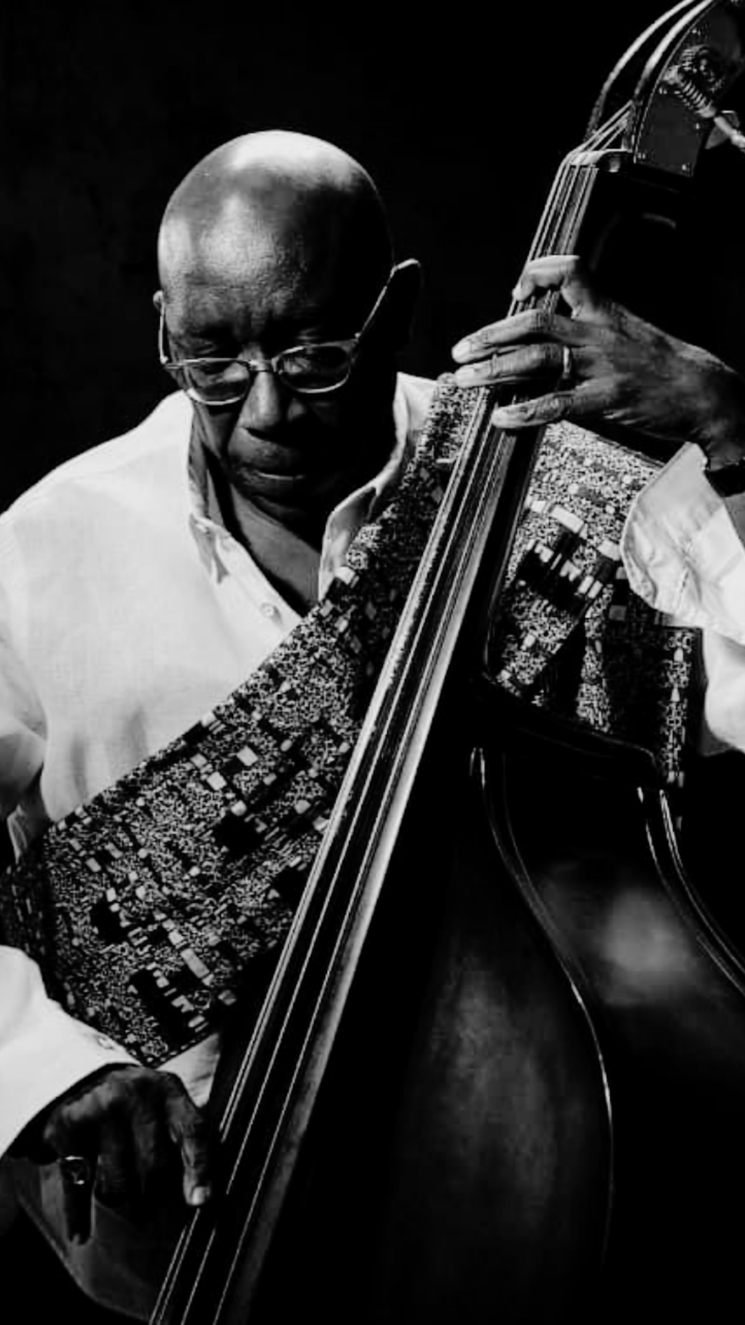 Black and white photo of a man playing an upright bass, wearing glasses and a white shirt, with a focused expression.
