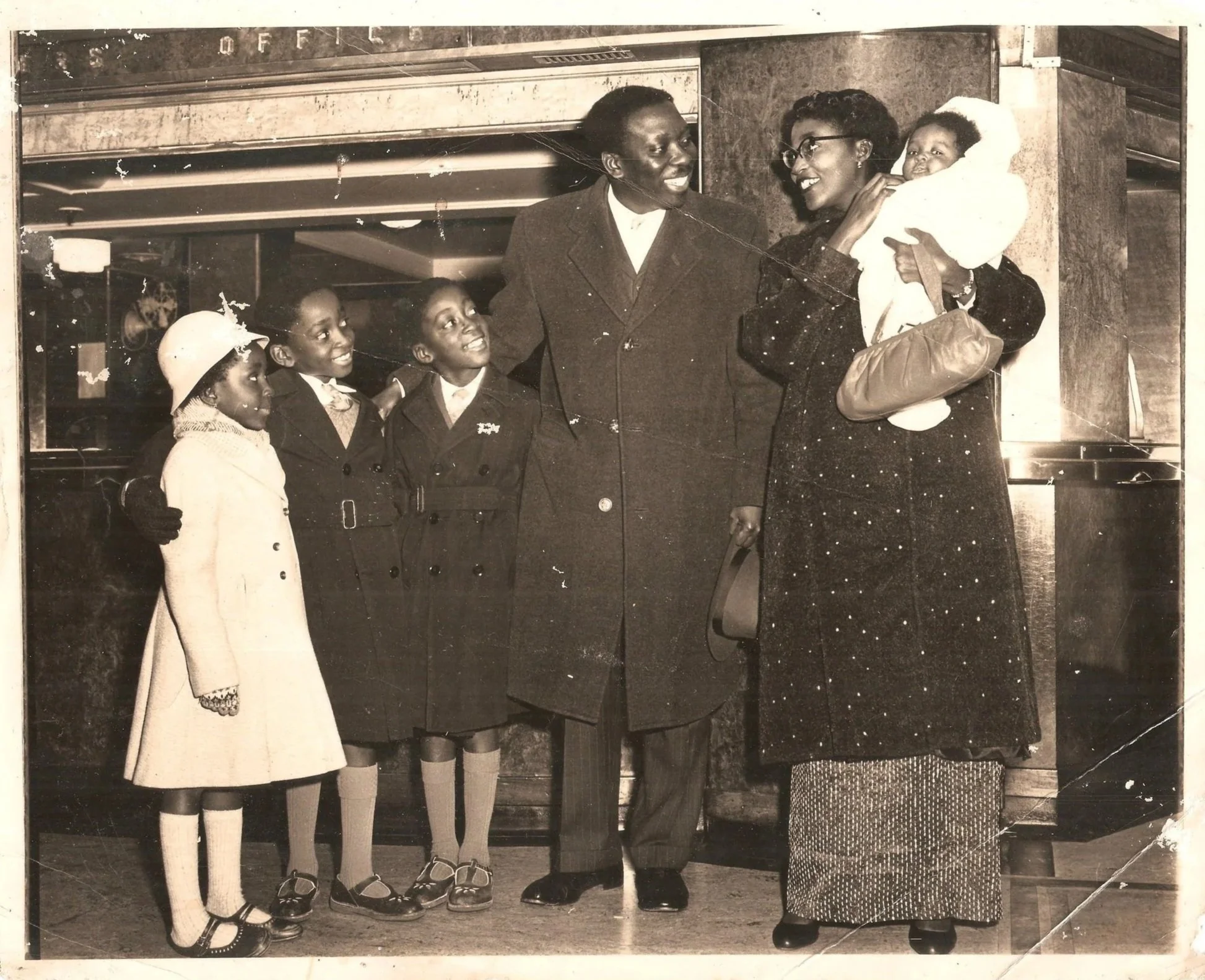 A black-and-white photo of a woman holding a baby, smiling at a man with three young children standing in front of her, in front of a building or establishment.
