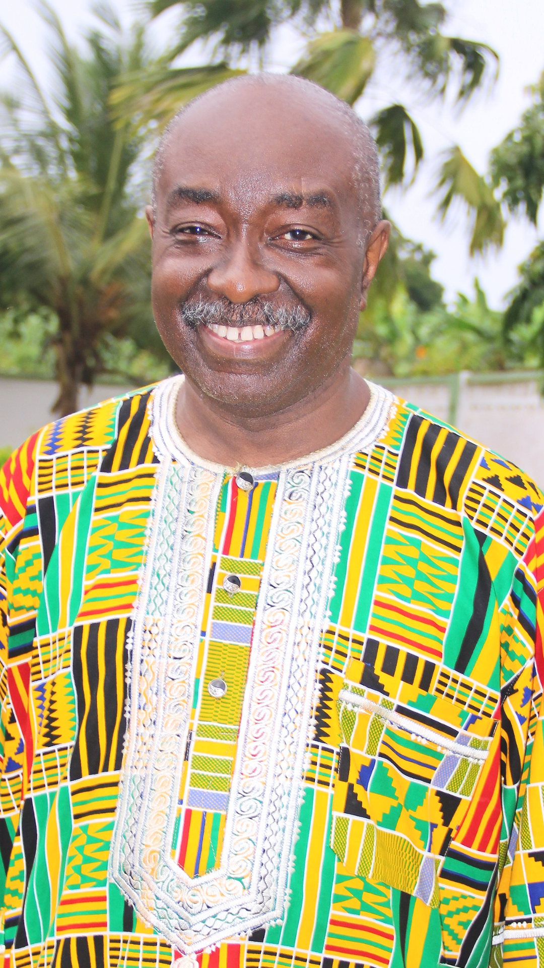 Smiling man wearing traditional African dashiki with colorful geometric patterns, outdoors with tropical trees in background.