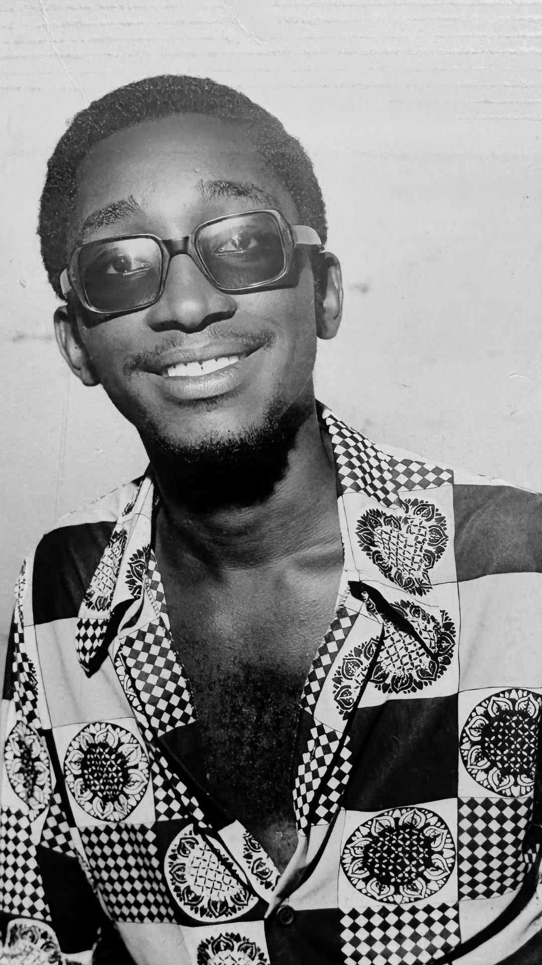 Teenage boy smiling, wearing glasses, and a patterned shirt, against a plain wall background.