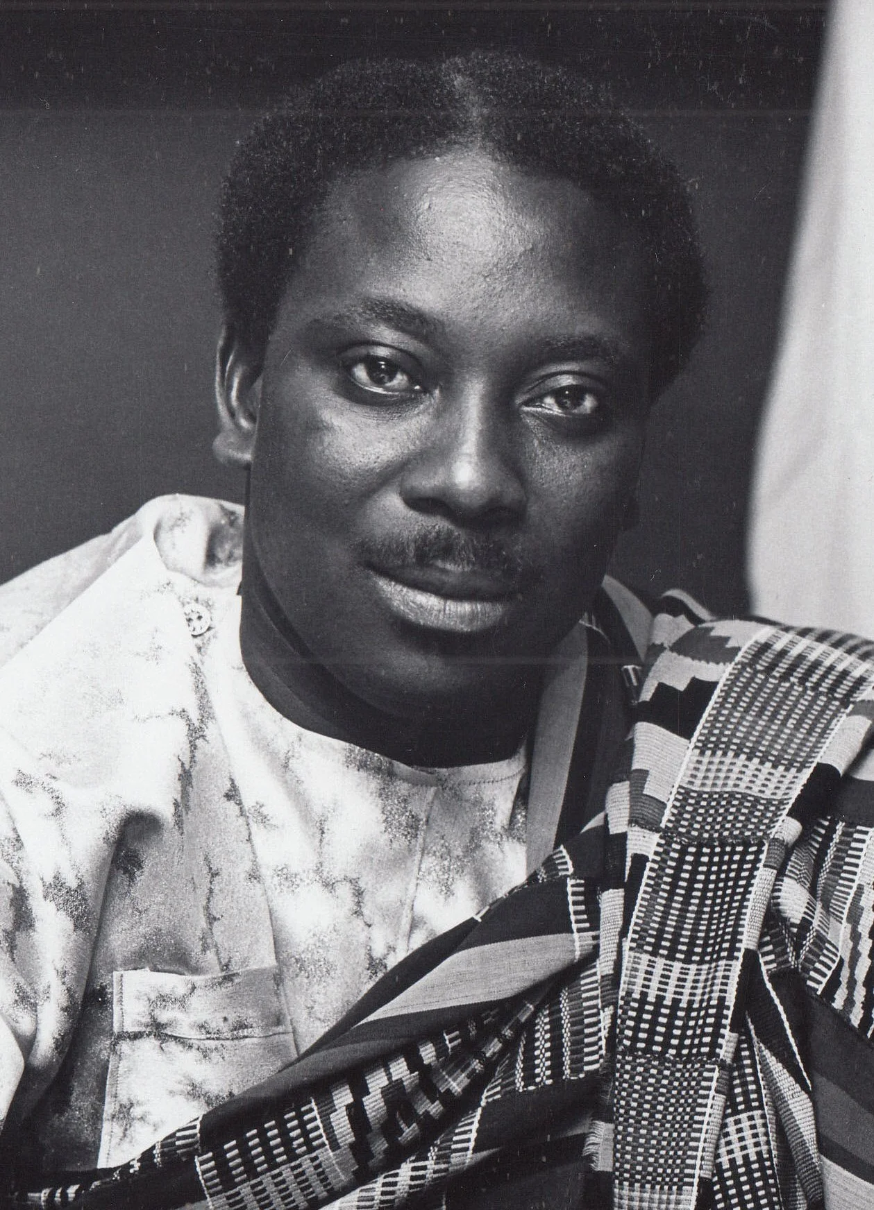 A black-and-white portrait of a man with short hair, wearing a kente a looking directly at the camera.