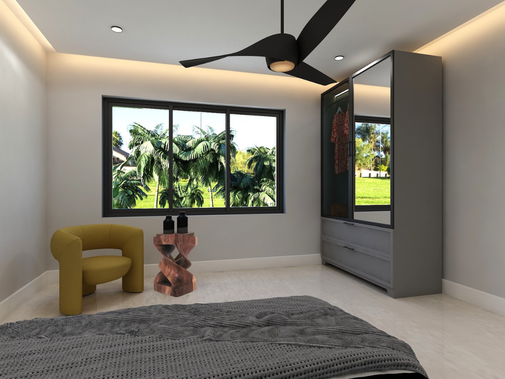 A modern bedroom with a large window showing a view of green trees and grass outside. The room has a ceiling fan, a yellow upholstered chair, a unique side table, a gray wardrobe with a mirror, and a dark gray blanket on the bed.