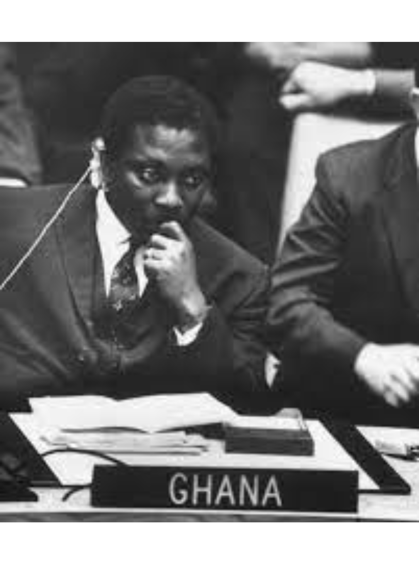 Black and white photo of a man in professional attire, sitting at a desk, with a sign that reads 'Ghana' in front of him.