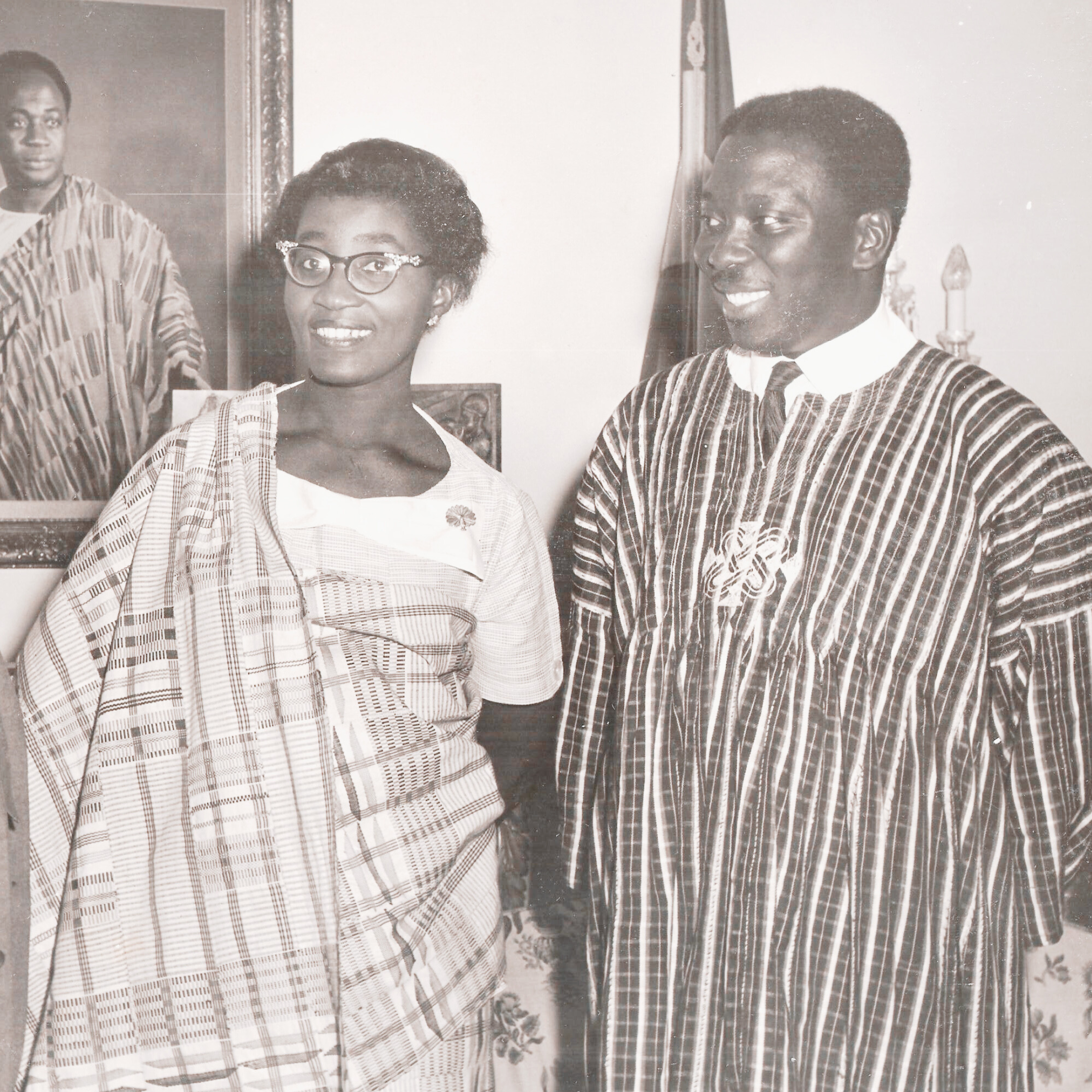Black and white photo of the Honorable Alex Quaison-Sackey and his wife, Elsie Quaison-Sackey  in traditional African clothing, standing indoors with a portrait of Kwame Nkrumah in the background.