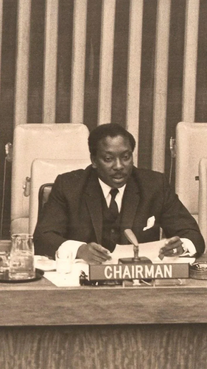 Black man in a suit sitting at a table with a microphone, holding a piece of paper, with a sign that reads "Chairman" in front of him, in a formal meeting room.