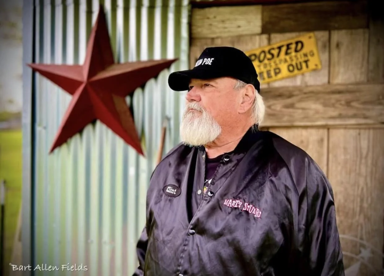 An elderly man with a white beard and mustache wearing a black cap and black satin jacket, standing outdoors near a rustic wooden wall and a corrugated metal sheet. A red star decoration is mounted on the wall behind him, and there is a yellow sign that reads "POSTED No Trespassing Keep Out."