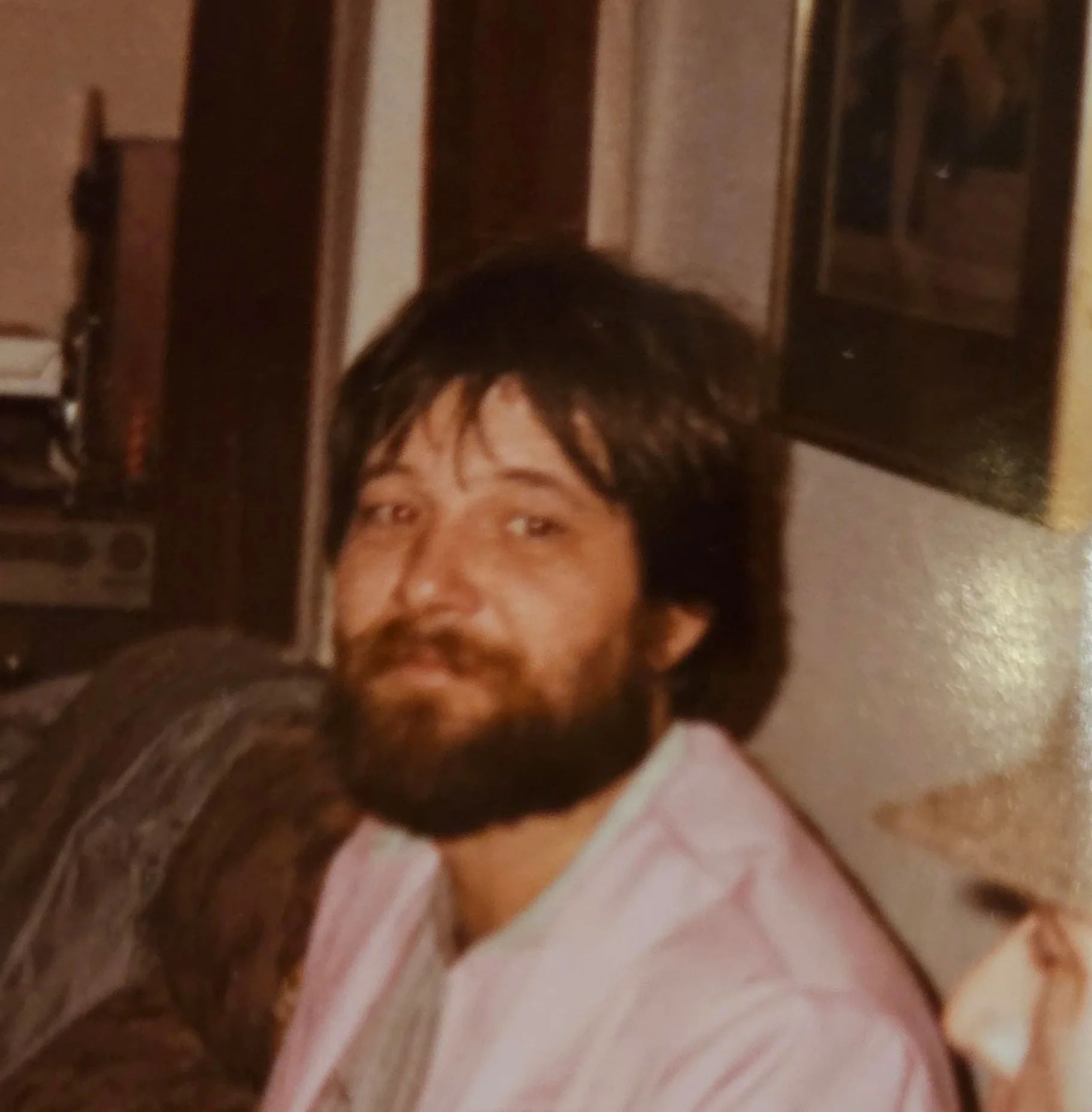 A man with dark hair and a beard, sitting indoors with a dark wooden cabinet and a framed picture in the background.