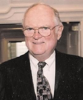 A middle-aged man with glasses, wearing a suit and a patterned tie, smiling in an indoor setting.