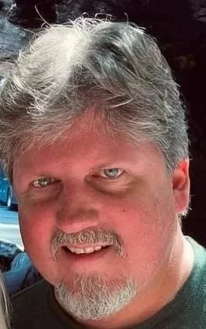Close-up of a man with gray hair and a beard, smiling outdoors.