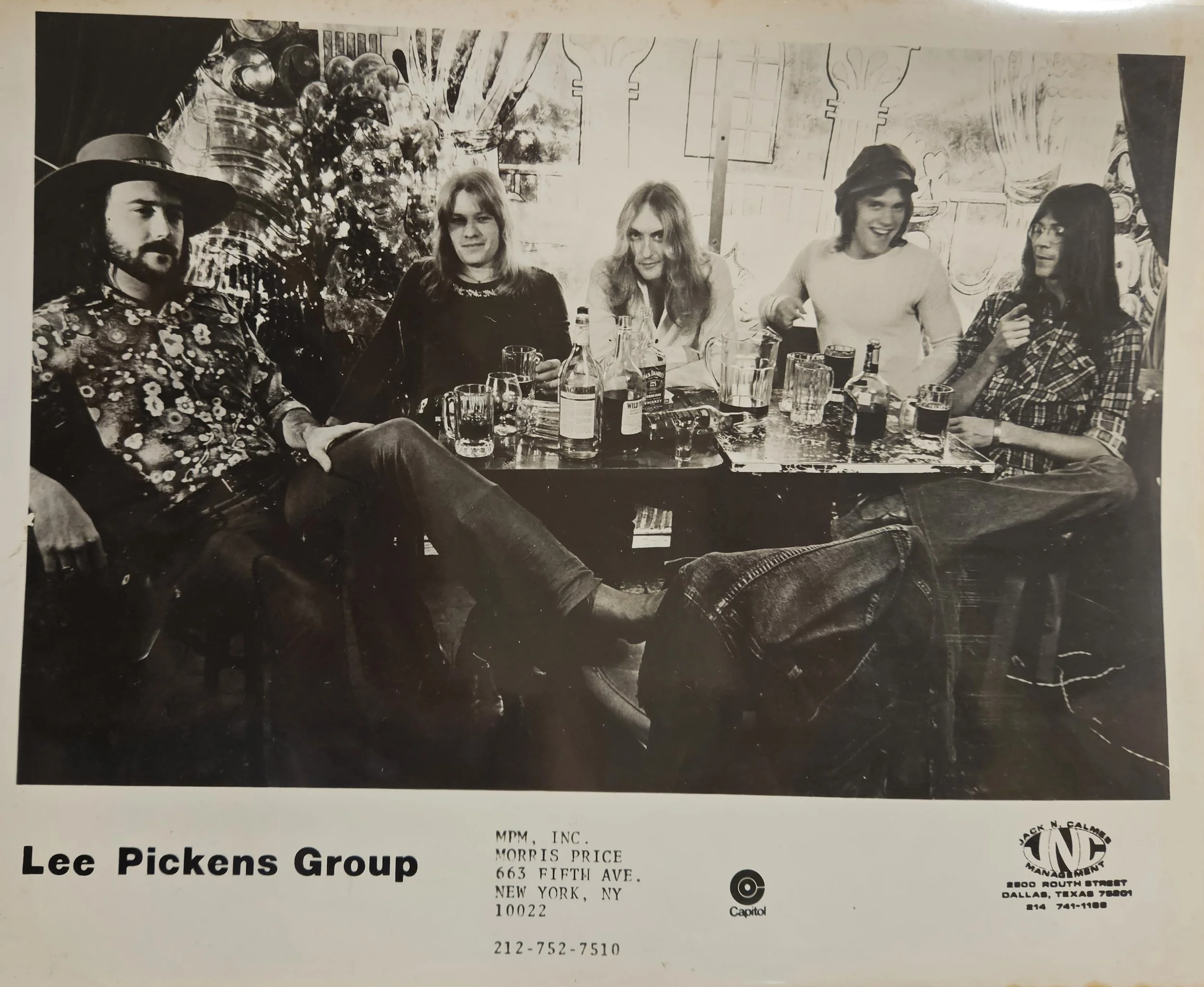 Black and white photo of the Lee Pickens Group, a group of five people sitting at a bar or restaurant table with drinks in front of them, smiling and enjoying each other's company, with a decorative mural in the background.
