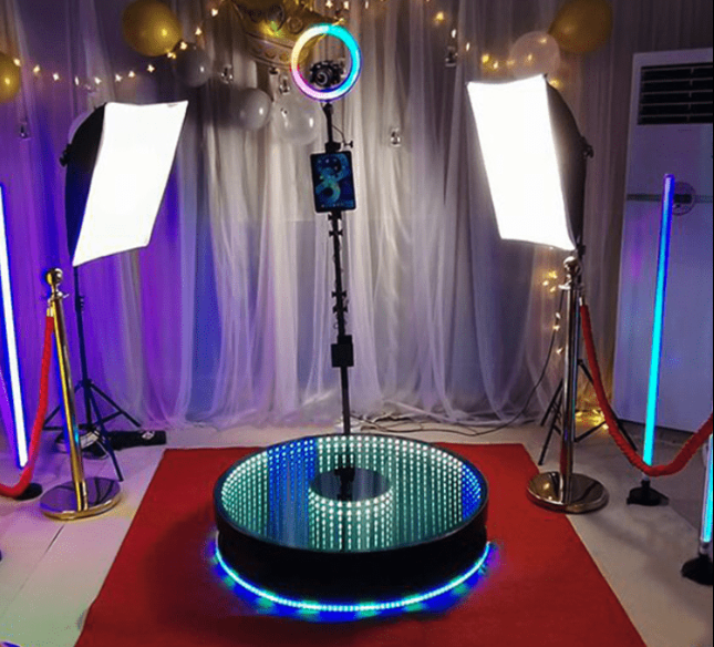 A photo booth setup with a rotating mirror ball, illuminated by two softbox lights, and a ring light with a colorful halo. There are red velvet ropes, a red carpet, and a large mirror with LED lights on the floor.