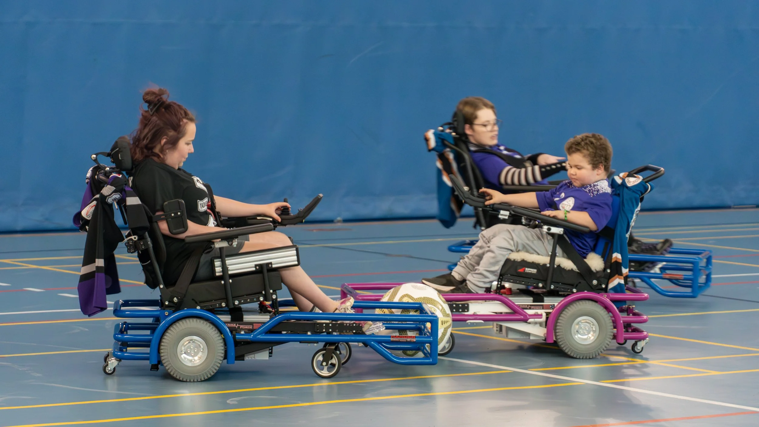 Two children in wheelchairs playing soccer indoors with a ball, one girl in a black shirt and one boy in a blue shirt and gray pants, on a gymnasium floor with colored lines.