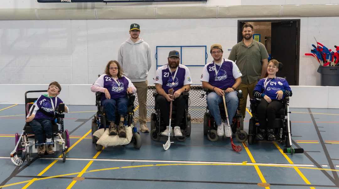 Group of five children in wheelchairs and two adult men standing behind them, inside an indoor sports facility with a hockey net and equipment in the background.