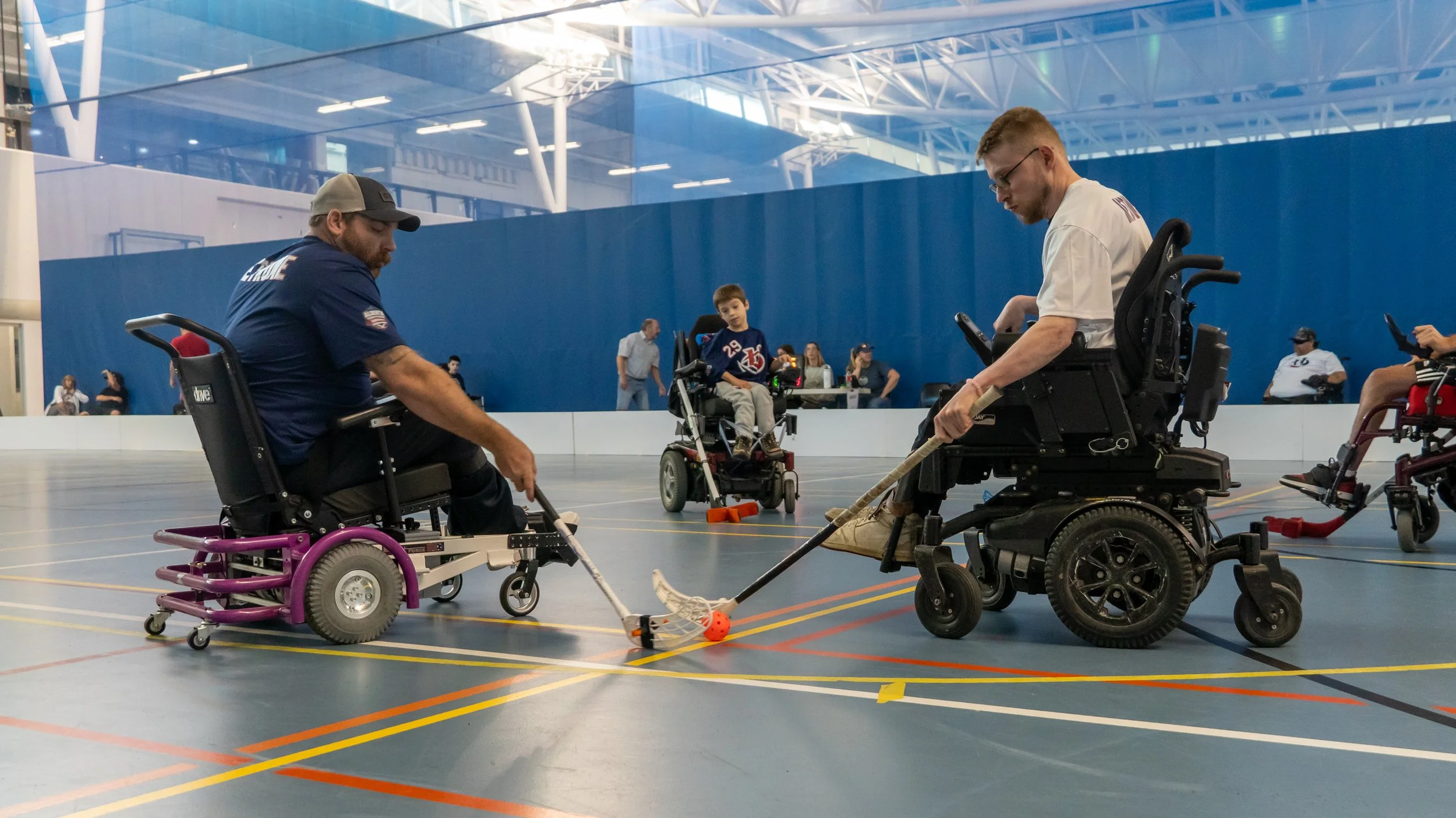 Two individuals in motorized wheelchairs playing wheelchair hockey with a small orange ball inside a large indoor sports facility, with a young boy and other spectators in the background.