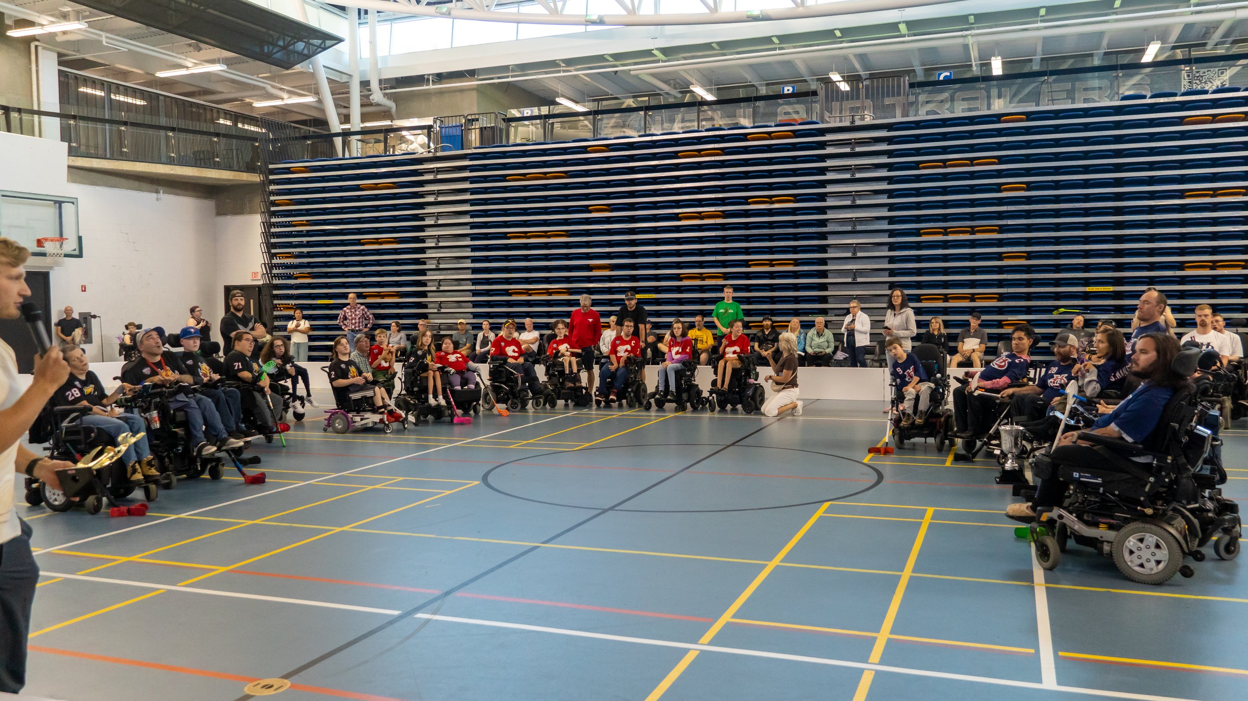 A large indoor gymnasium with people in wheelchairs sitting in a semi-circle, some holding sports equipment. A person is speaking with a microphone to the left, and others are standing or sitting in the background. The bleachers are mostly empty, and the gym floor has painted lines for sports.