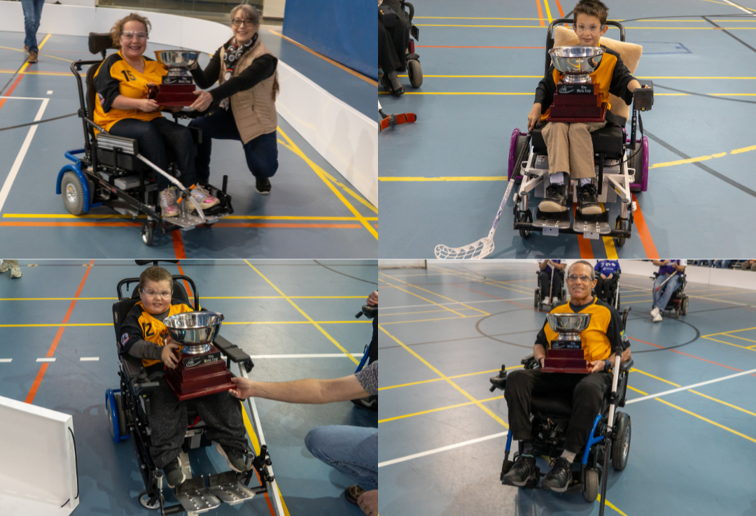 Four children in wheelchairs holding trophies at an indoor sports event, with two women presenting awards and other people in the background.