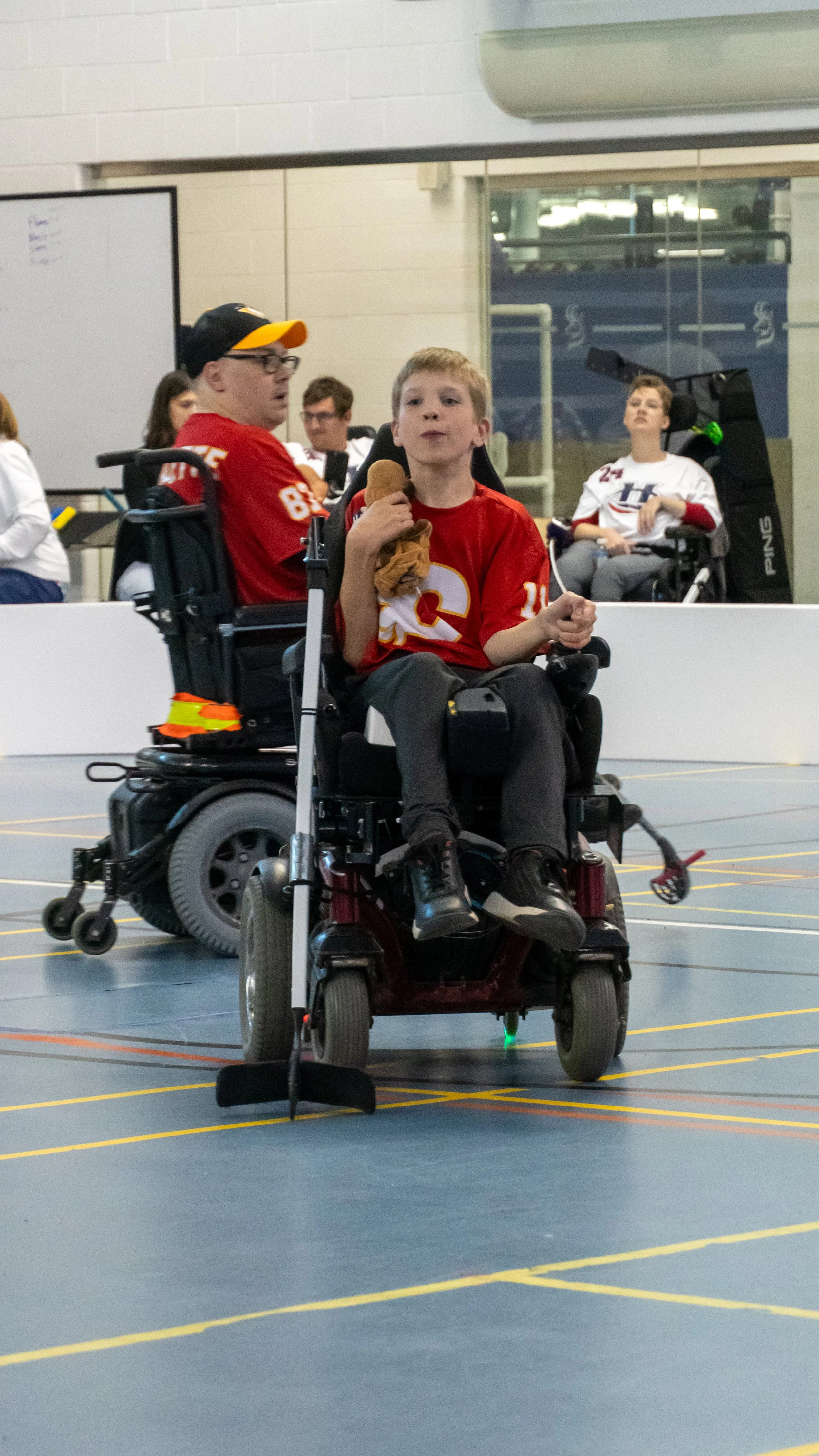 A young boy in a red sports jersey sitting in a motorized wheelchair, holding a stuffed toy and a snack, with other people in wheelchairs in the background.