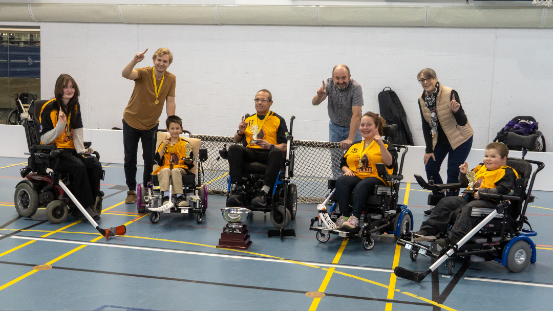 Group of six children in wheelchairs with two adults in a gym, holding a hockey stick and trophies, celebrating a sporting event.