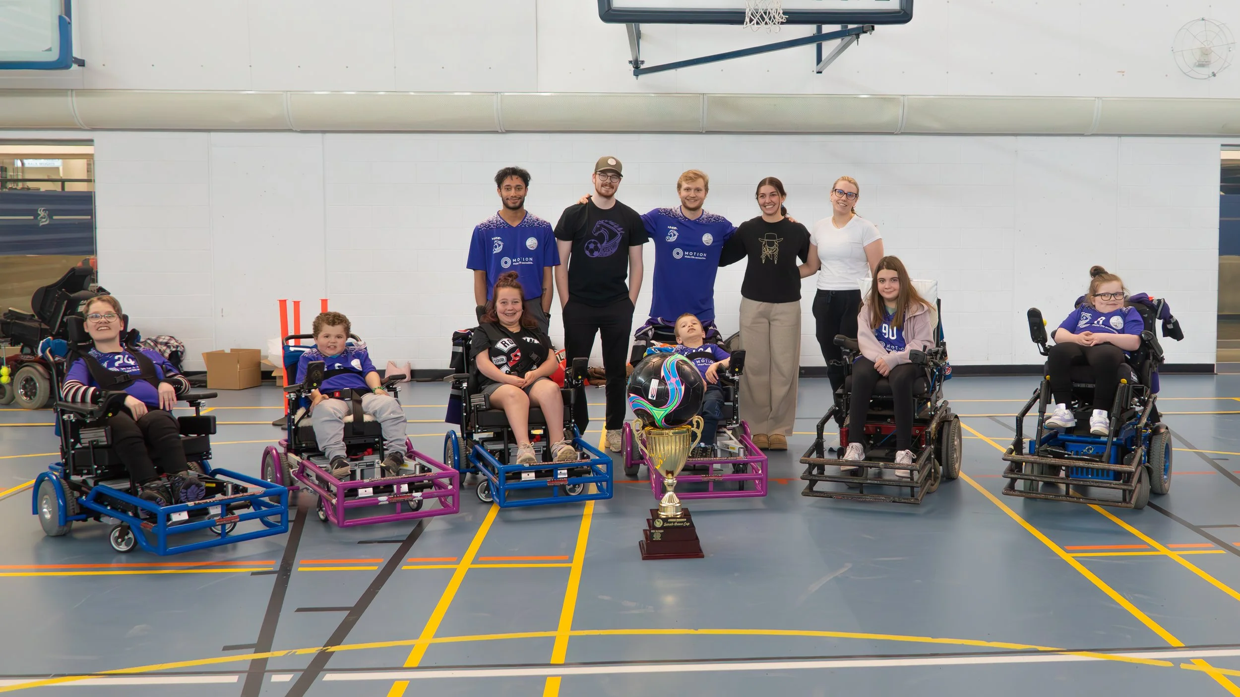 Group of seven children in wheelchairs and three young adults in a gymnasium, with a trophy and a helmet in front, celebrating after a sports event.