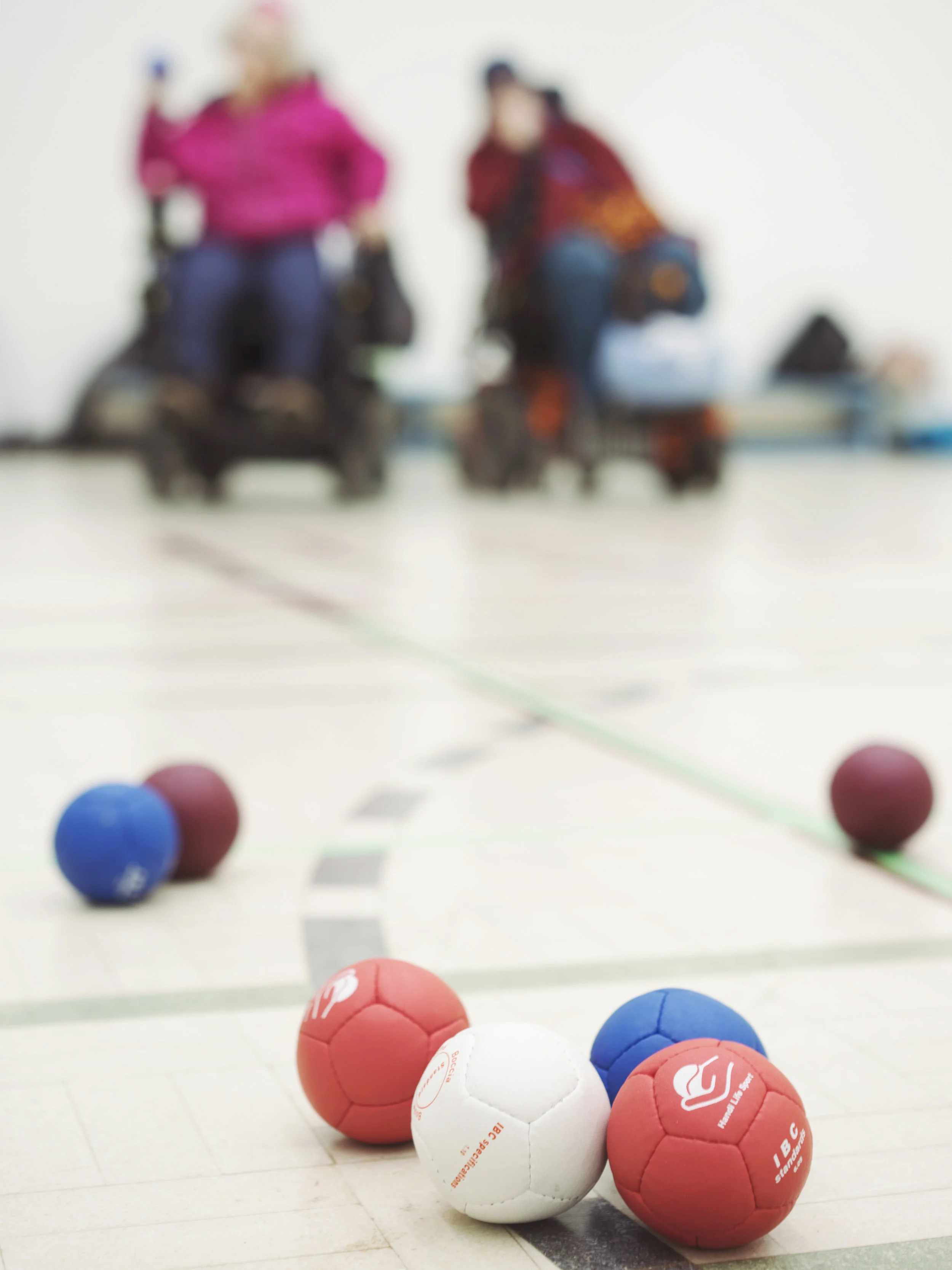 Specialized indoor sports balls on a court, with two players in wheelchairs in the background, blurred.